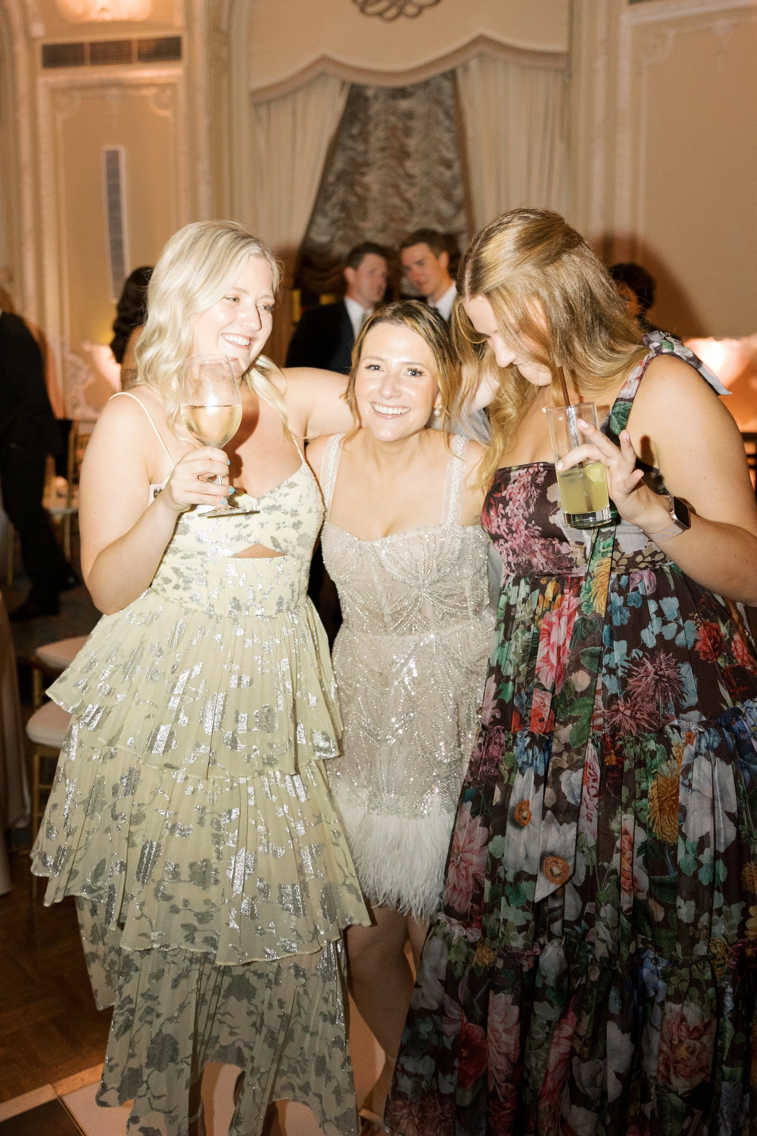 Group of three women at a formal event, smiling and holding drinks, with other guests in the background.