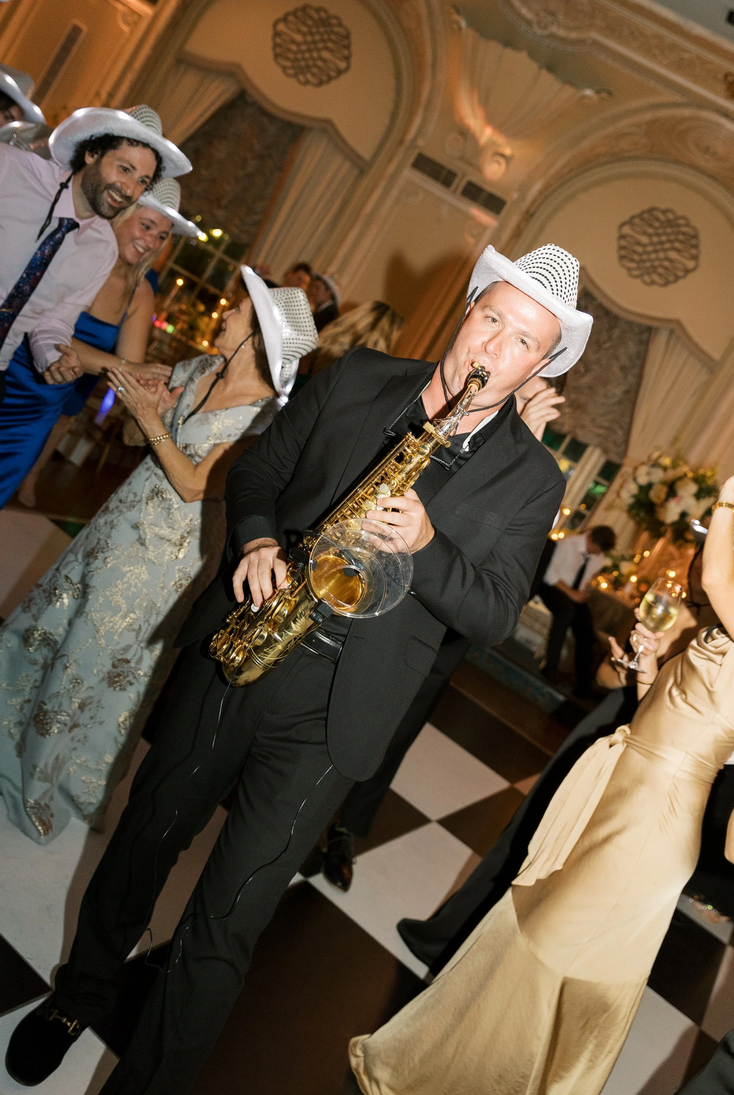Man in black suit playing saxophone at a celebration, wearing a white hat, with guests dancing and socializing in an ornate ballroom.