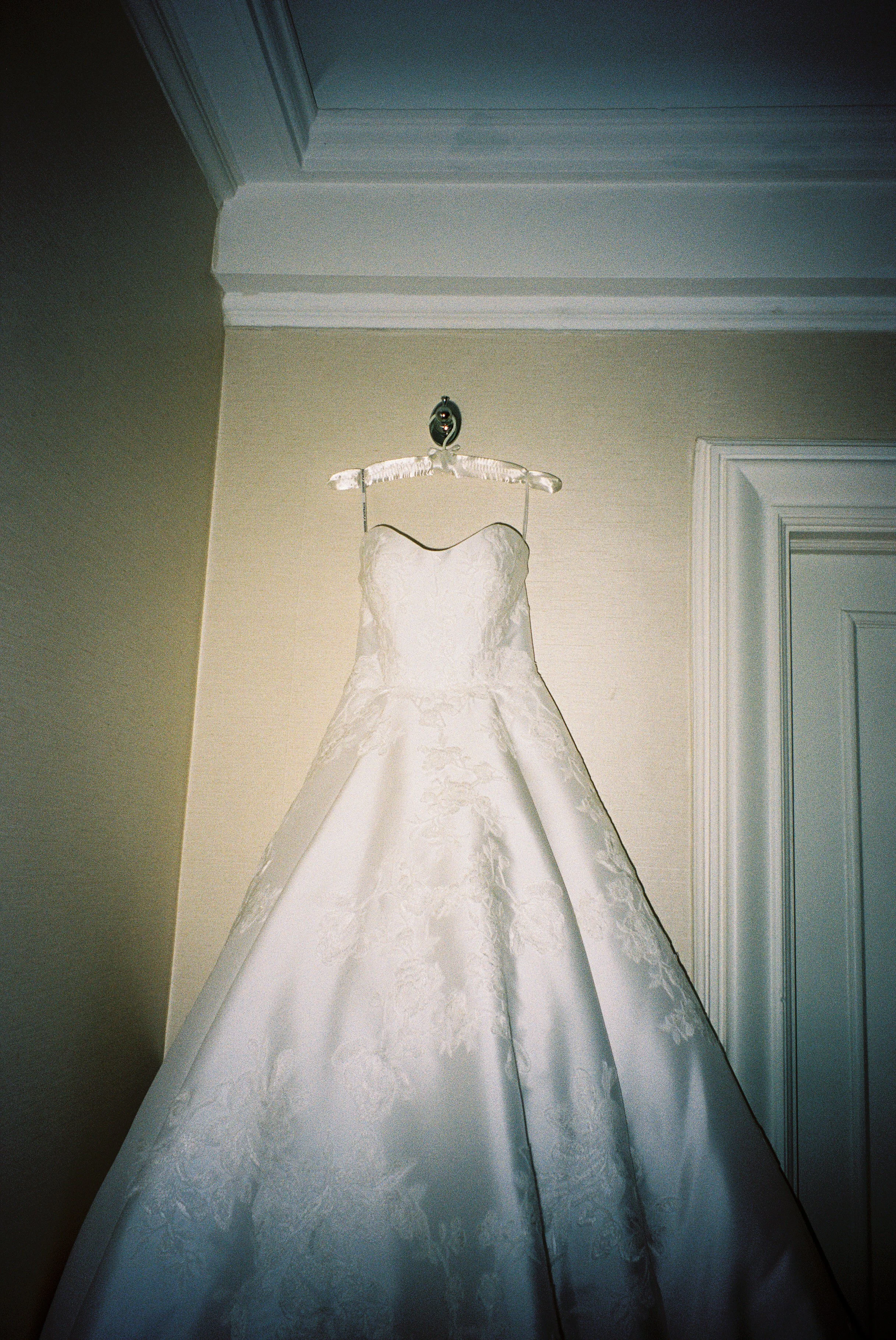 A white wedding dress hanging on a hanger against a beige wall with crown molding.