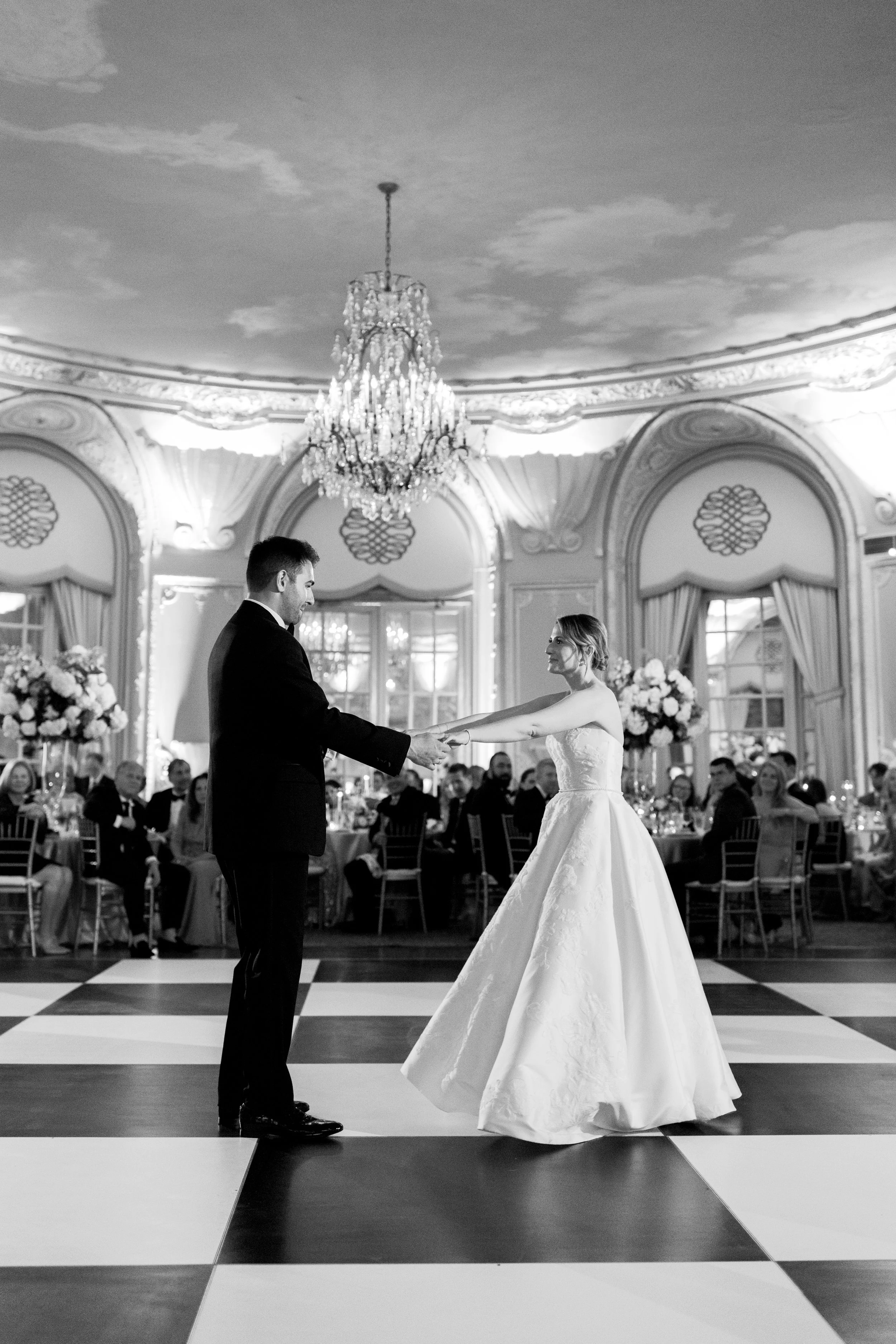 A bride and groom holding hands while dancing at their wedding reception in an elegant ballroom with chandeliers and large floral arrangements.