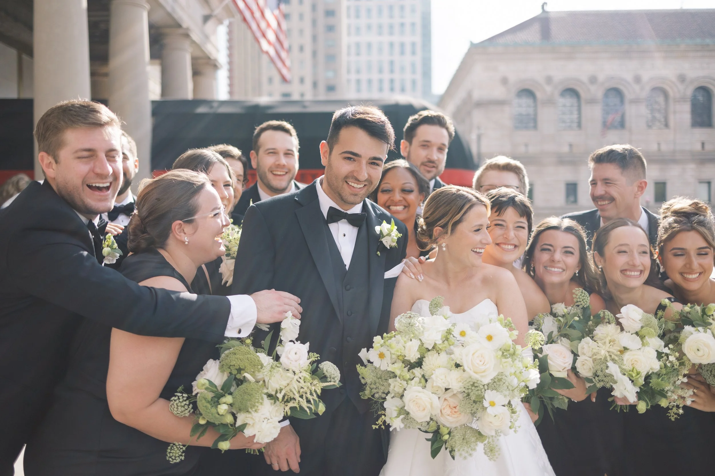 A bride and groom surrounded by a group of smiling wedding guests holding bouquets outside in an urban setting.