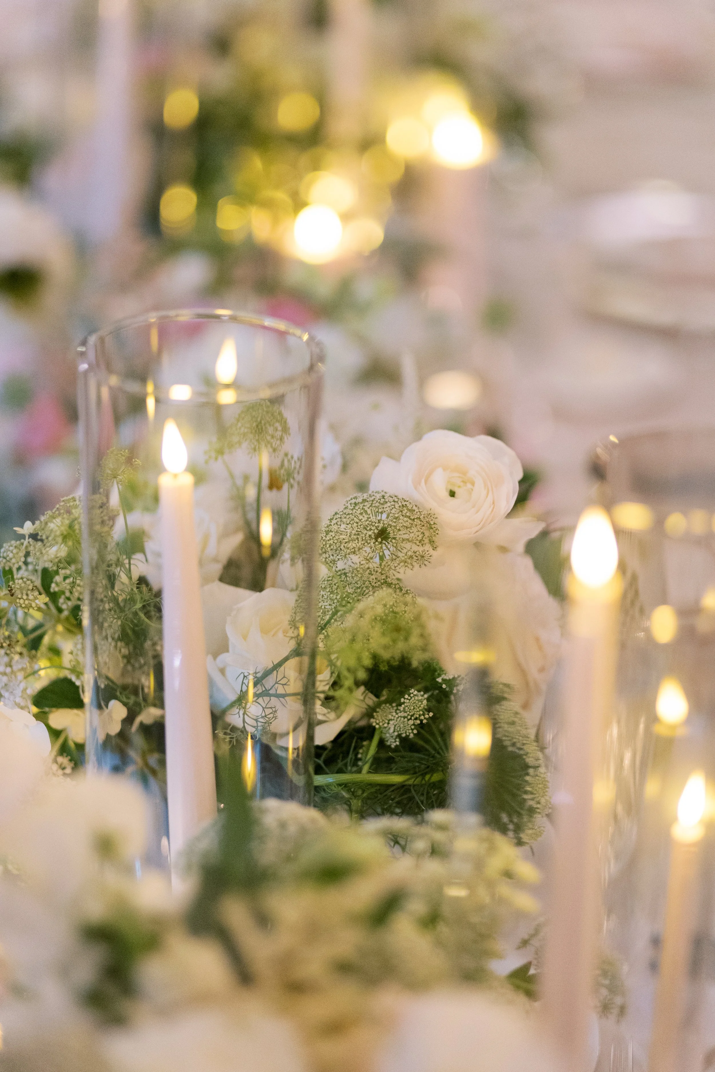 Elegant table centerpiece with white roses, greenery, and tall glass candles with flickering flames.