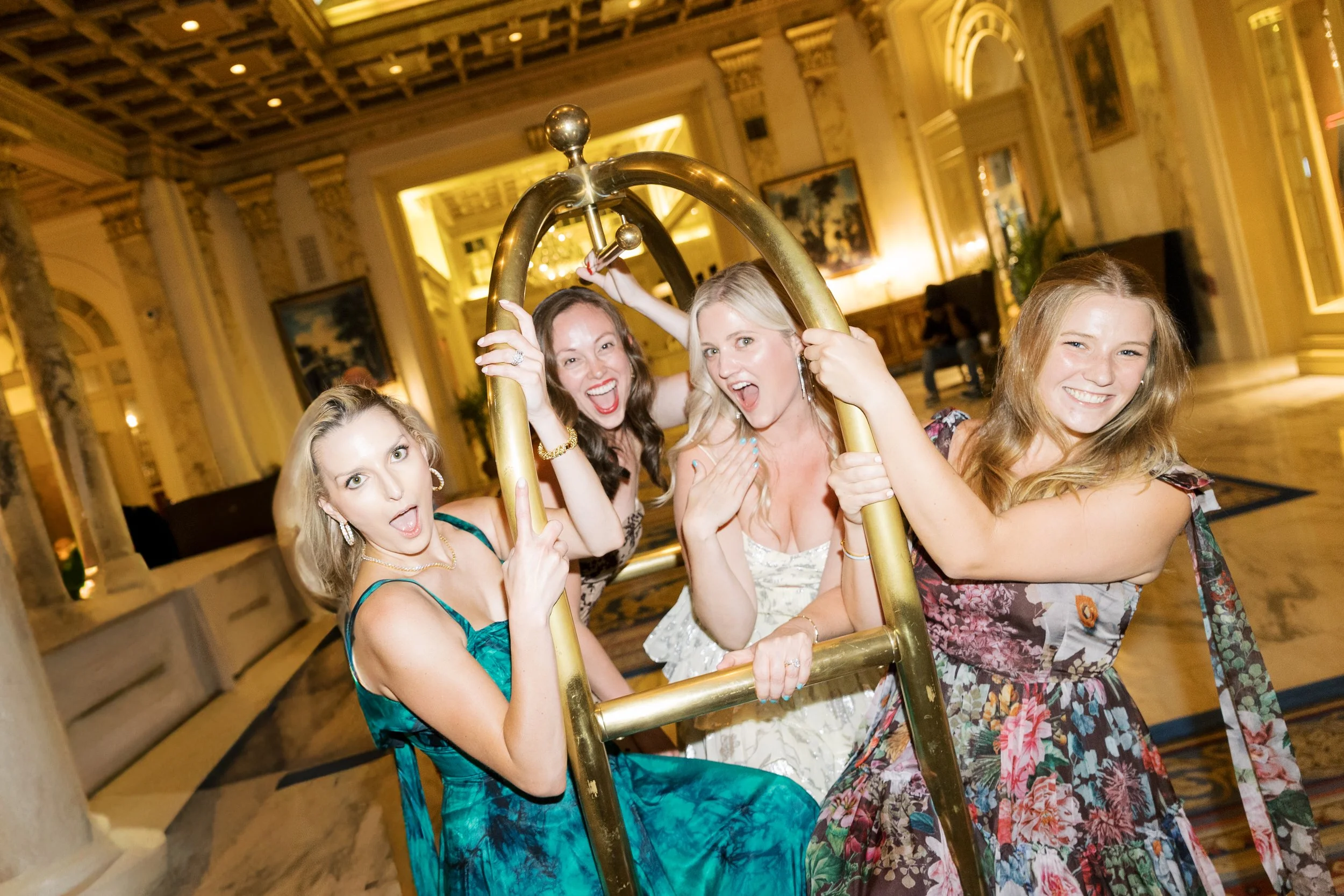 Four women in colorful dresses, smiling and having fun, seated on a brass luggage cart in an elegant lobby with marble floors, chandeliers, and framed artwork.