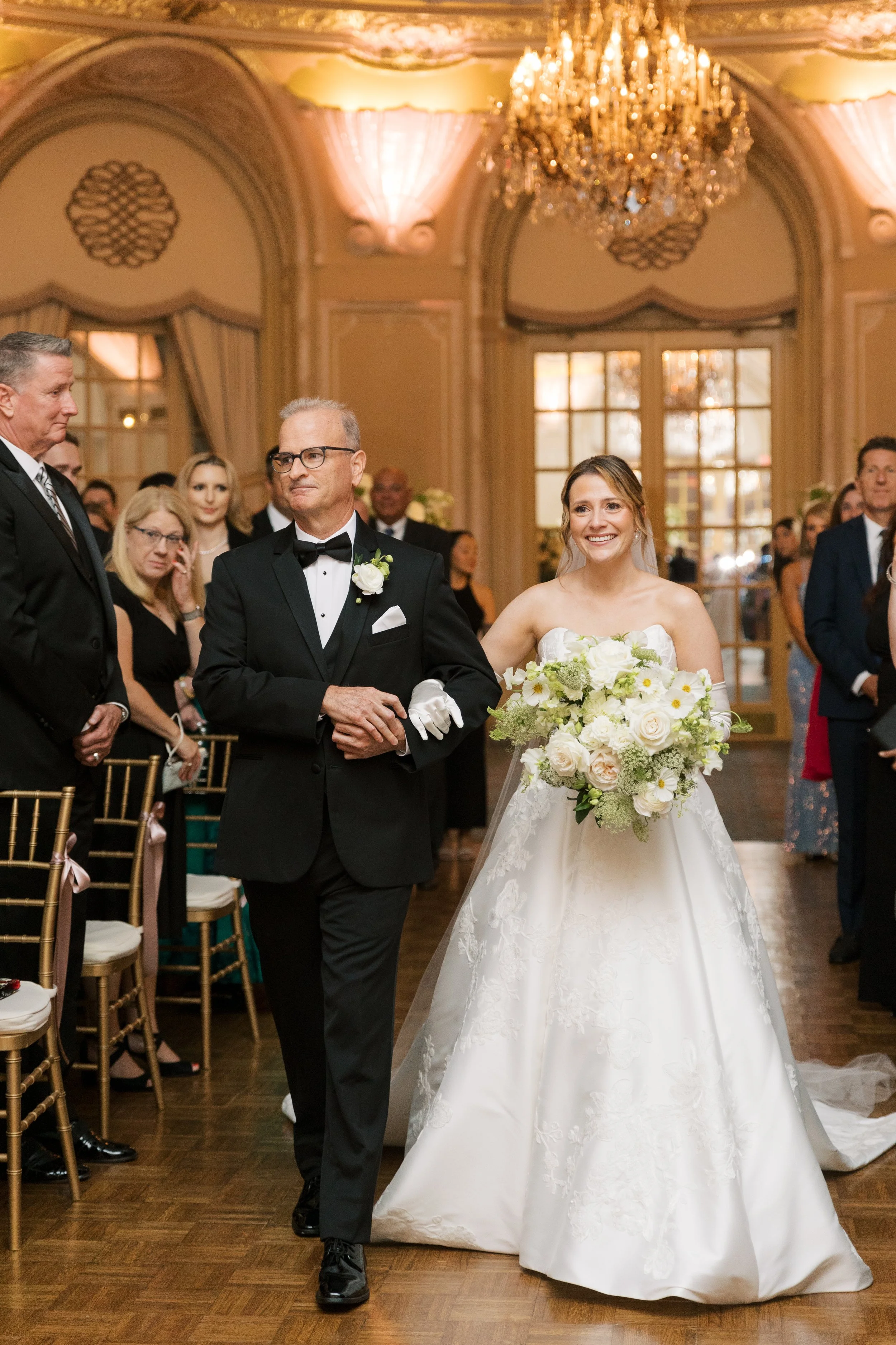 A bride walking down the aisle with her father at a wedding ceremony inside a grand, elegantly decorated hall with a chandelier.
