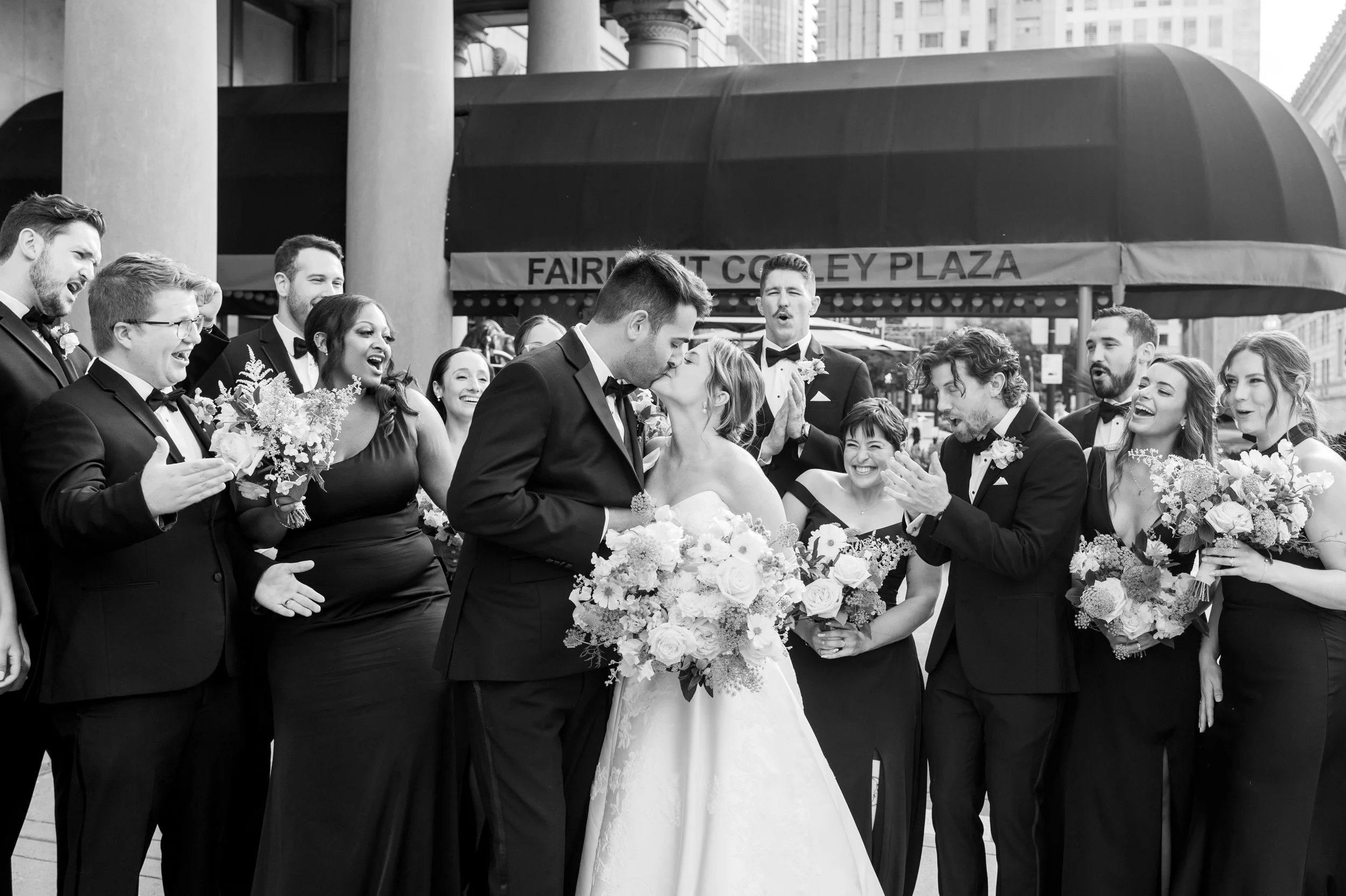 A black and white photo of a wedding scene taking place outdoors in front of a building with a sign that reads 'Fairmont Copley Plaza.' A newlywed couple is in the center, sharing a kiss while surrounded by smiling bridesmaids and groomsmen, some hol