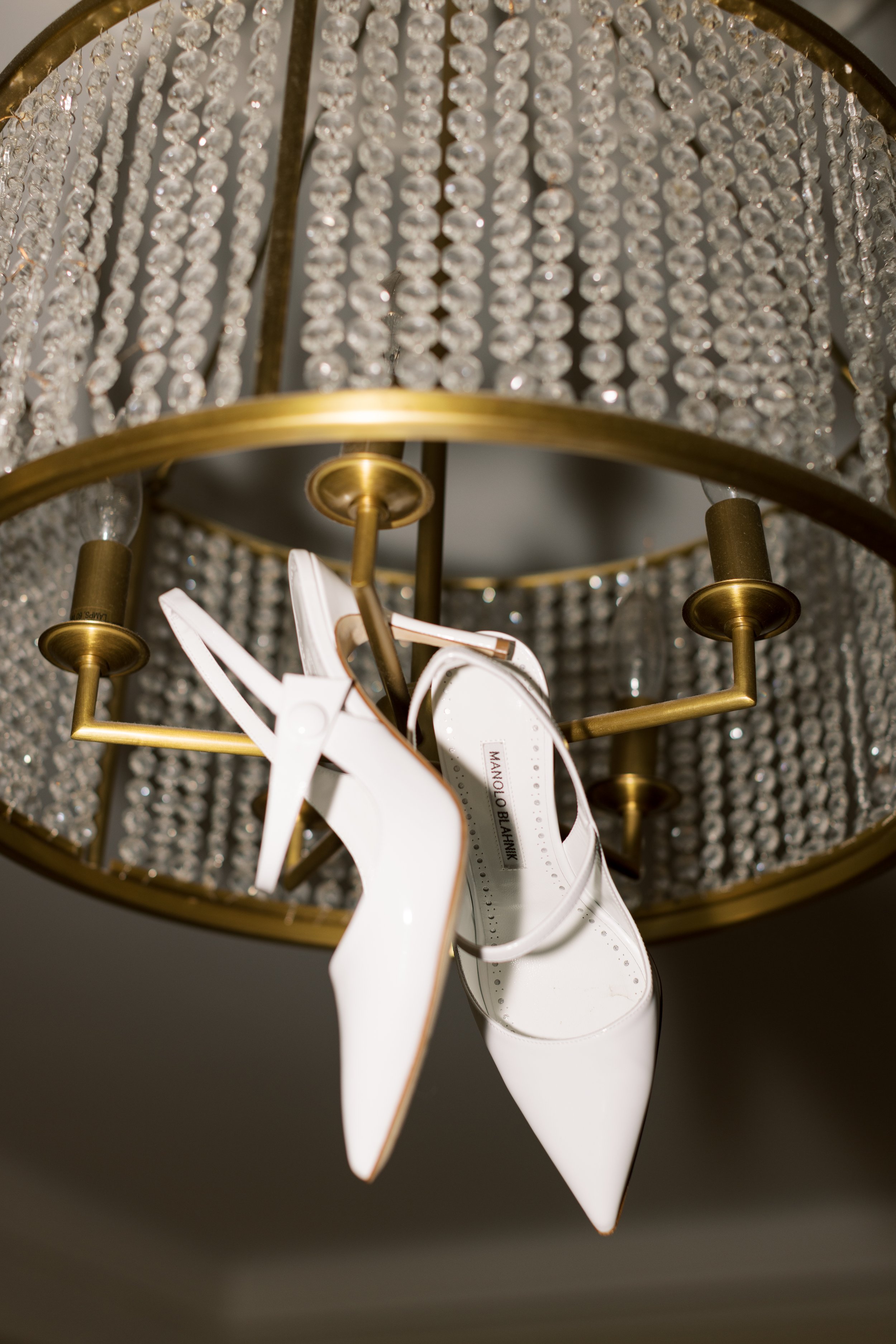 White high-heeled shoes hanging from a chandelier with crystal beads and gold accents.