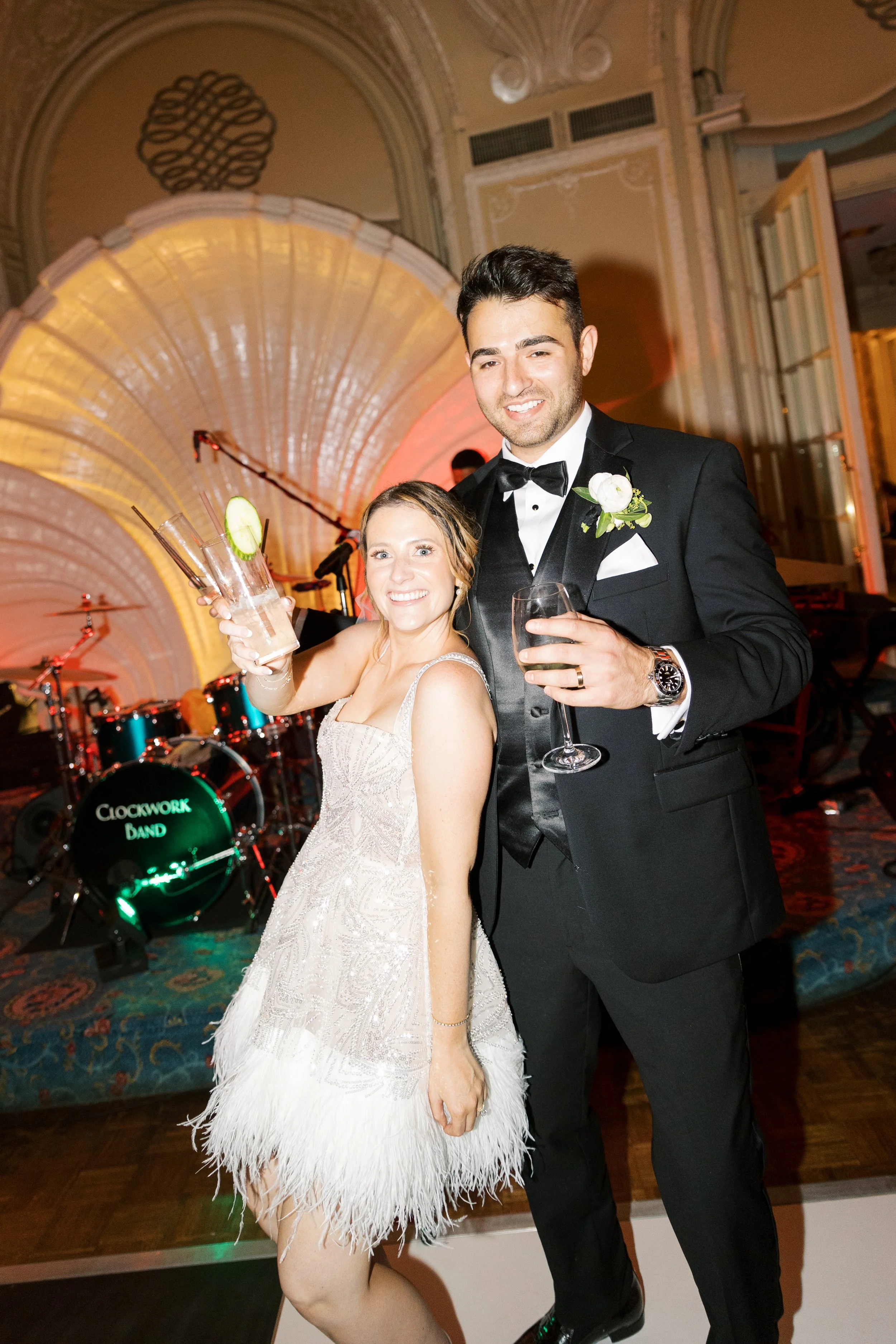 A bride and groom at their wedding reception, standing together and smiling, with the bride holding a drink with a cucumber slice and the groom holding a glass of red wine, in a decorated ballroom with a musical band in the background.