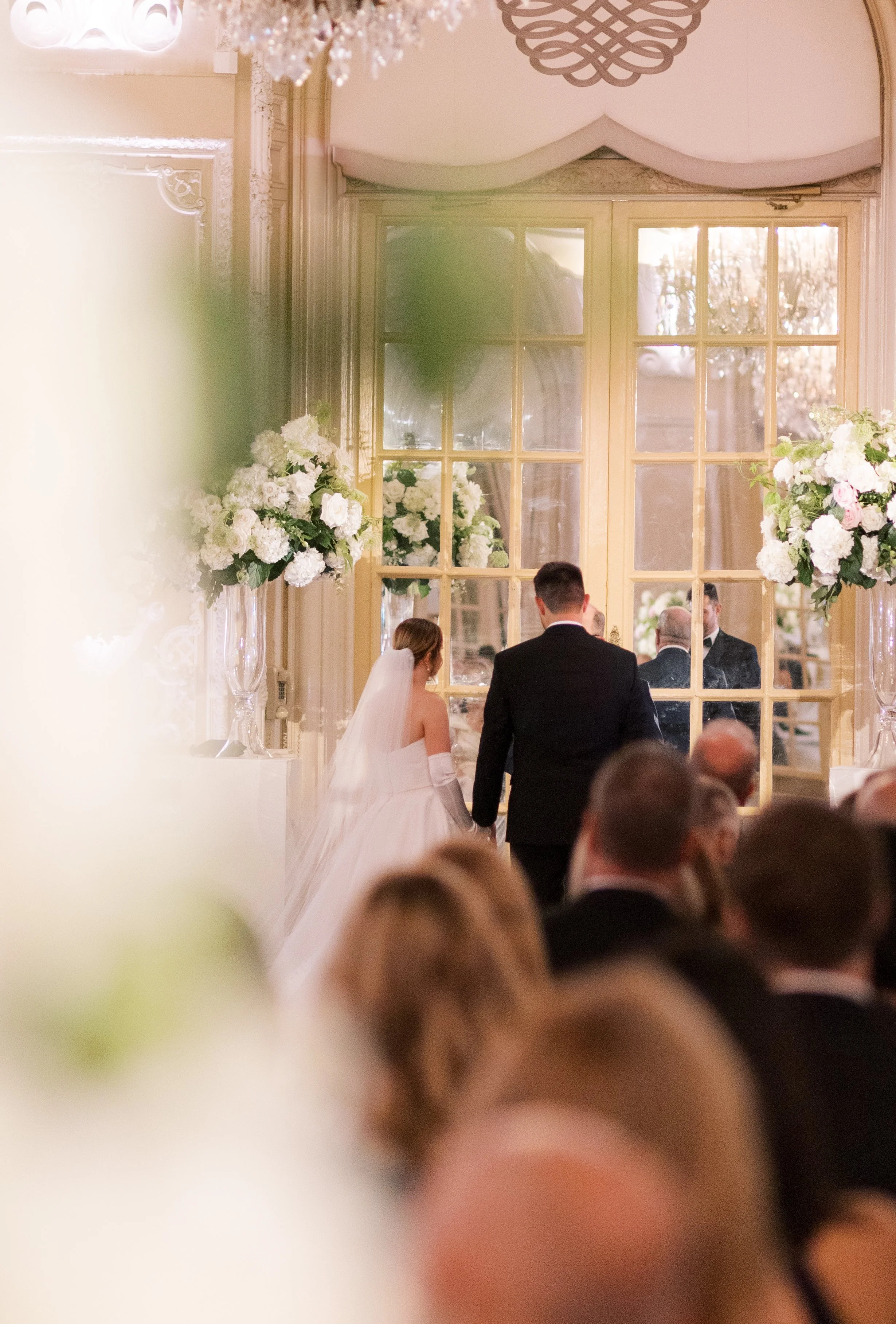 Bride and groom holding hands during their wedding ceremony, facing a large window with floral decorations, surrounded by seated guests.