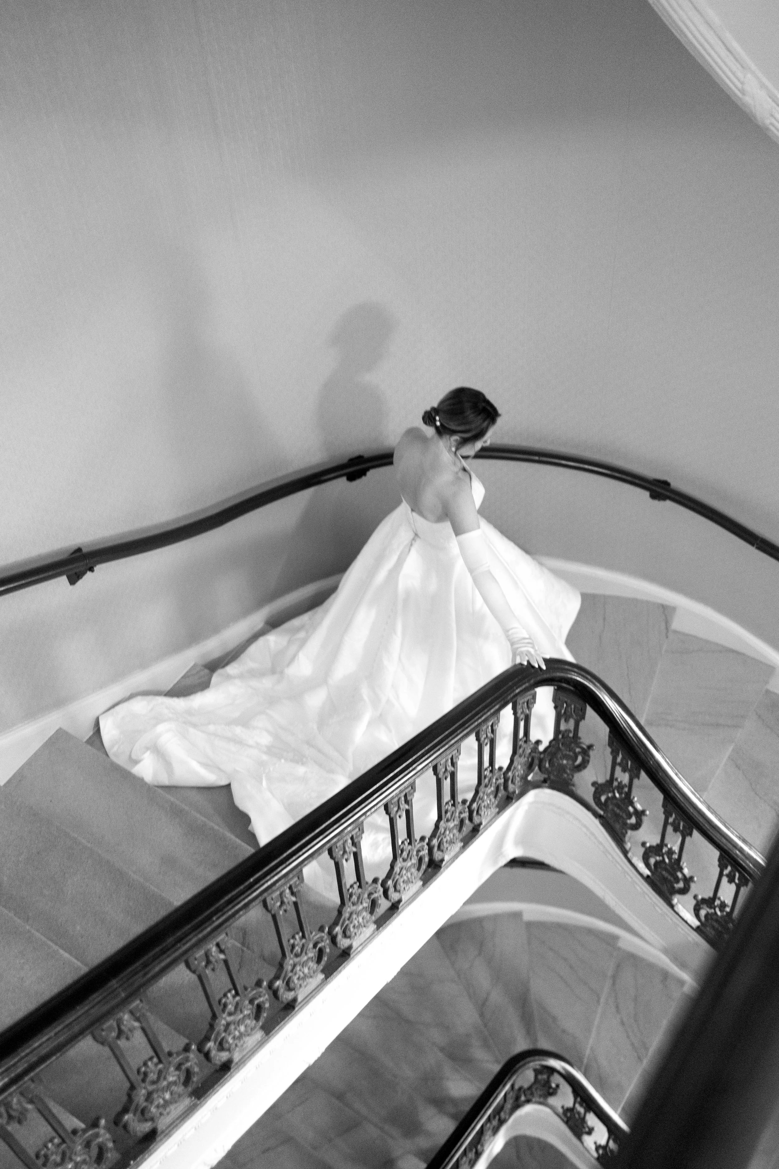 A woman in a wedding dress stands on a staircase, looking down, with her back to the camera and gloves on, illuminated by soft lighting.