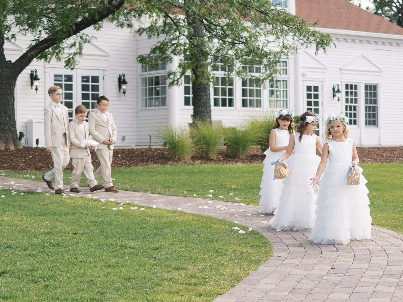 A group of three boys in light beige suits and three young girls in white dresses with floral headbands, walking on a brick pathway in front of a white house with large windows and green plants.