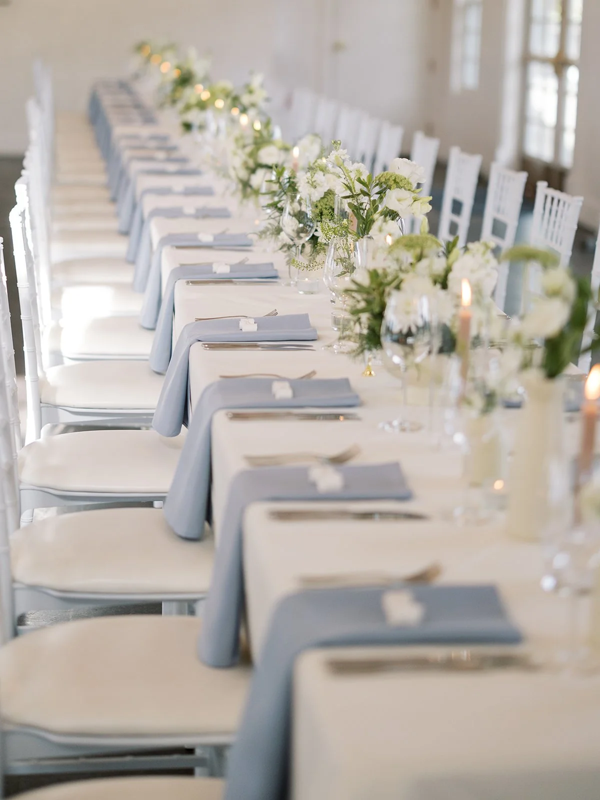 Elegant wedding or banquet table set with white tablecloths, folded gray napkins, floral centerpieces, wine glasses, candles, and white chairs in a bright room.