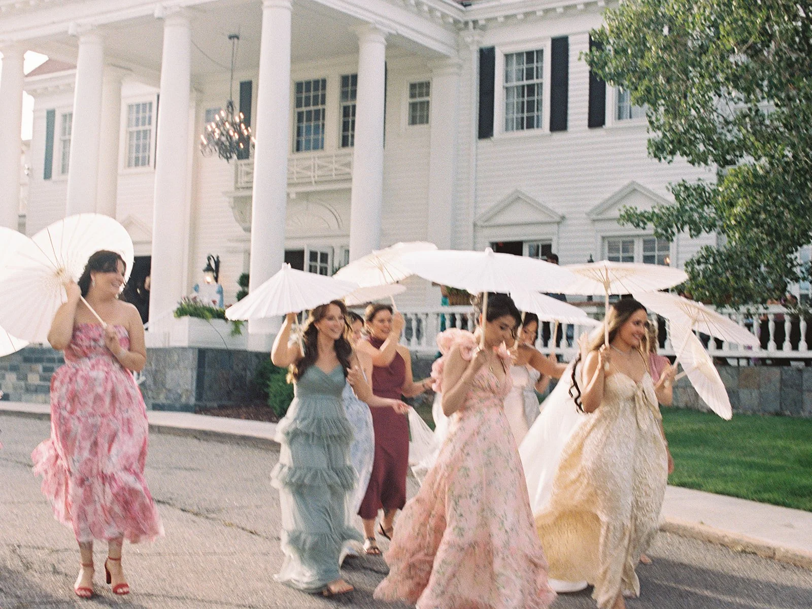 Group of women in dresses walking outdoors, holding white parasols, in front of a large white house with pillars.