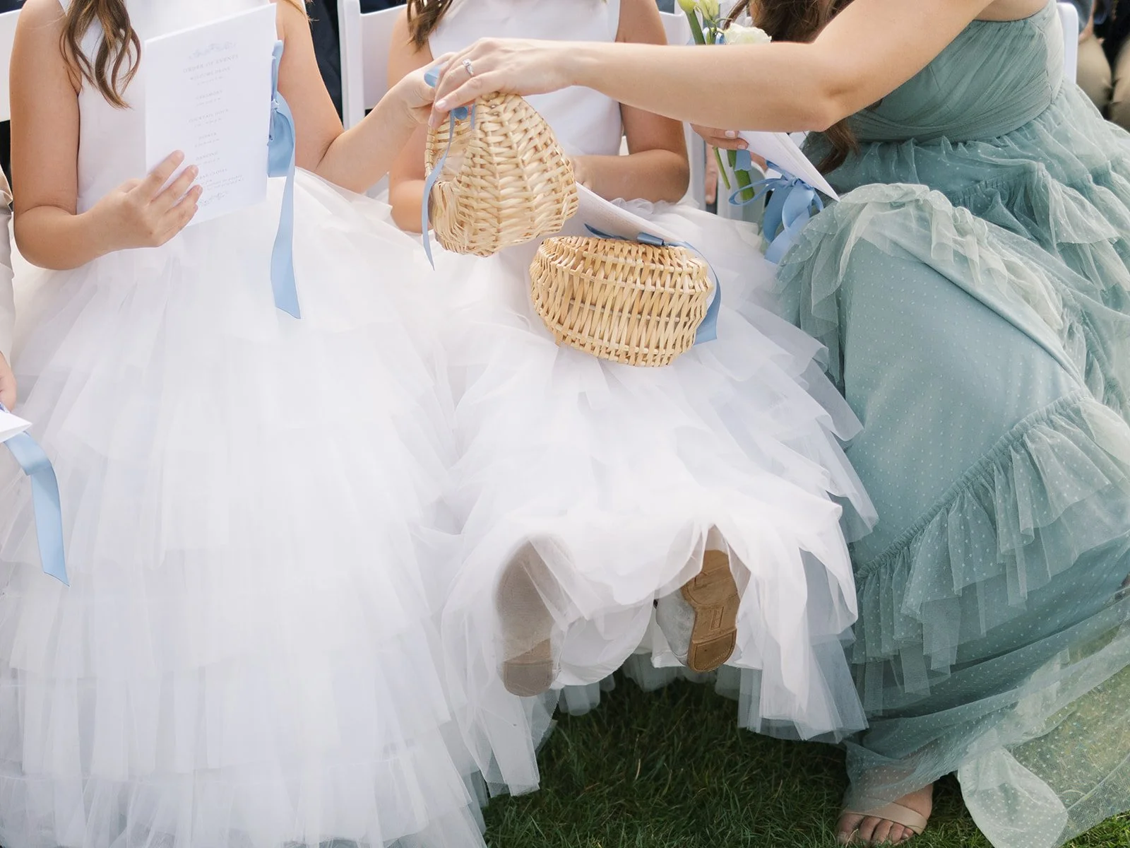 Children dressed in white and light blue dresses, holding papers, sitting at a table with woven baskets during a wedding ceremony.