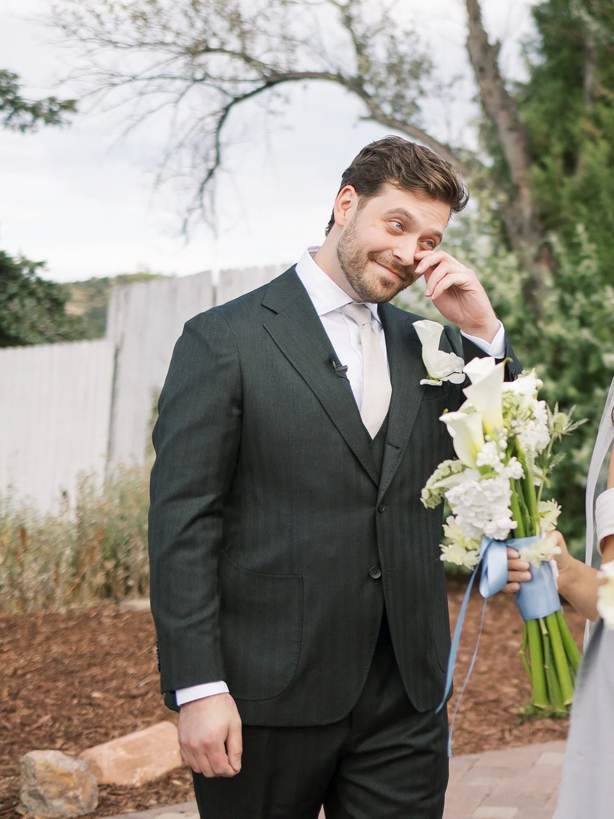 A man in a dark suit and tie, with a white boutonniere, is wiping his eye while smiling at a woman holding a bouquet of white flowers during an outdoor wedding.