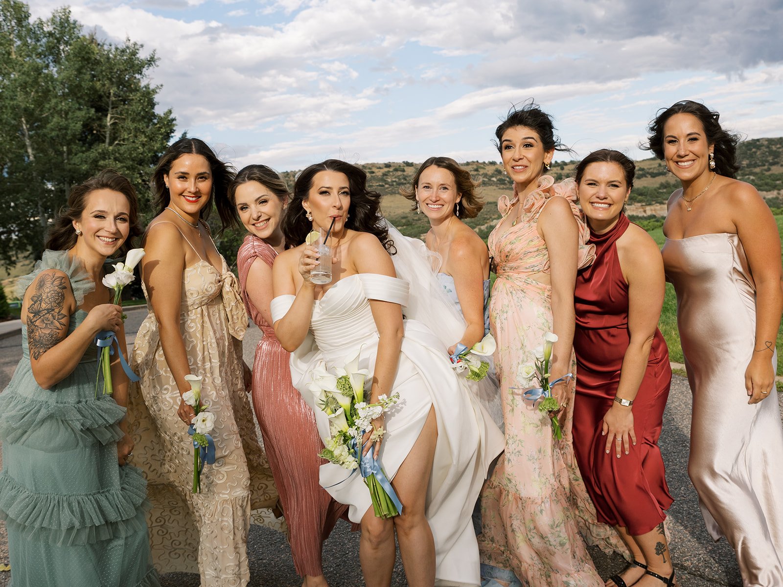 Group of women, including a bride, standing outdoors in a scenic landscape during a celebration, dressed in colorful dresses with flowers.
