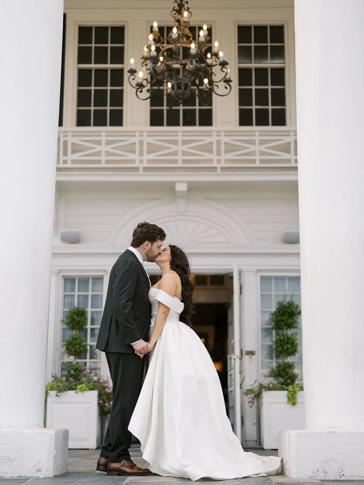 Bride and groom sharing a kiss in front of a white building with large windows, potted plants, and a chandelier inside.
