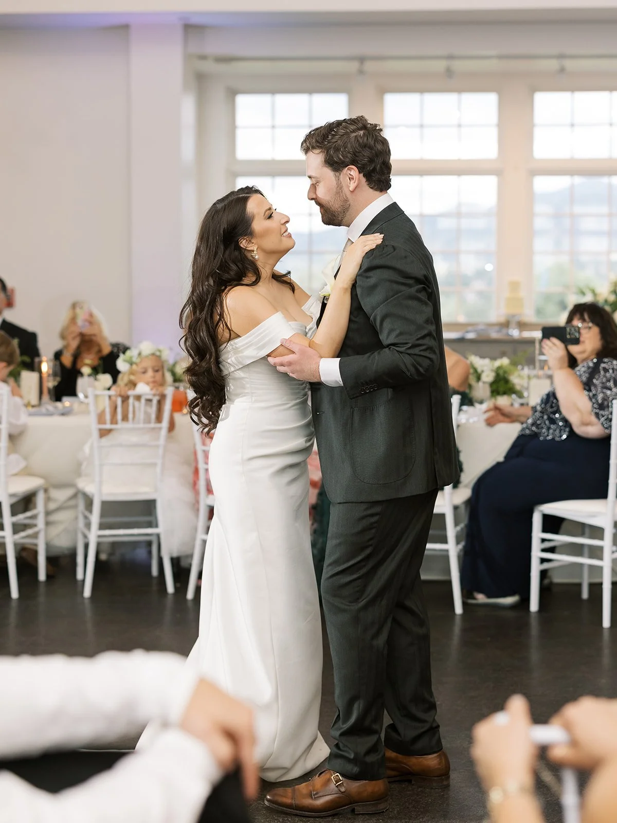 A bride and groom dancing in a wedding reception, with guests in the background taking photos and watching.