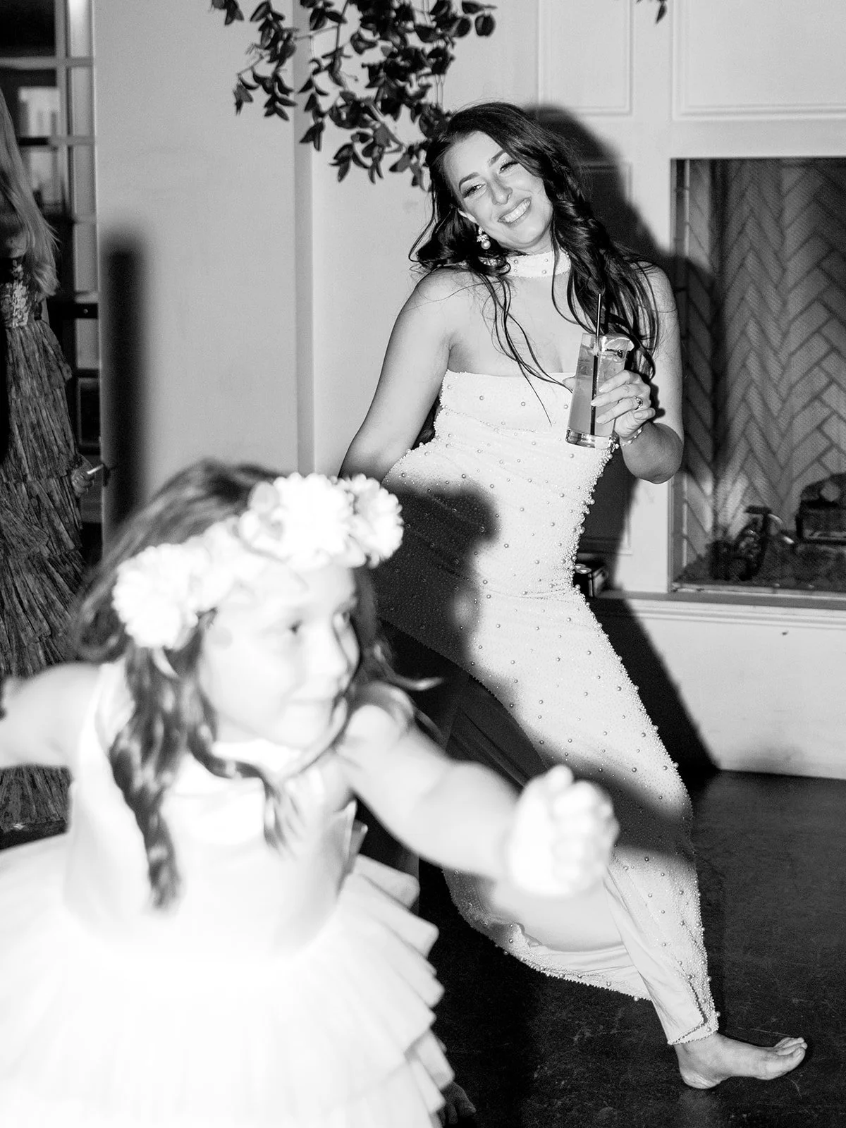 A woman in a white dress is smiling and holding a drink, dancing at a celebration. A young girl with a flower crown and light-colored dress is also dancing in the foreground.