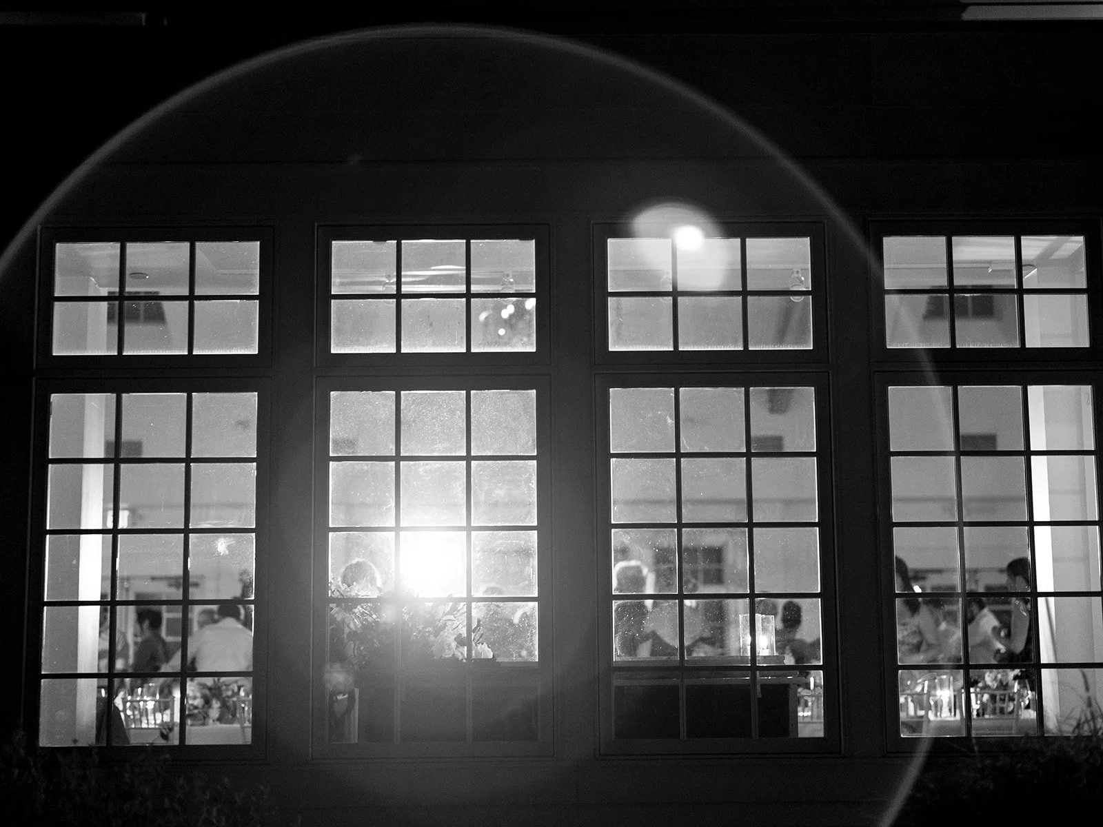 Black and white photo of a large window with multiple panes, showing a scene inside a well-lit venue with people gathered for an event or gathering.