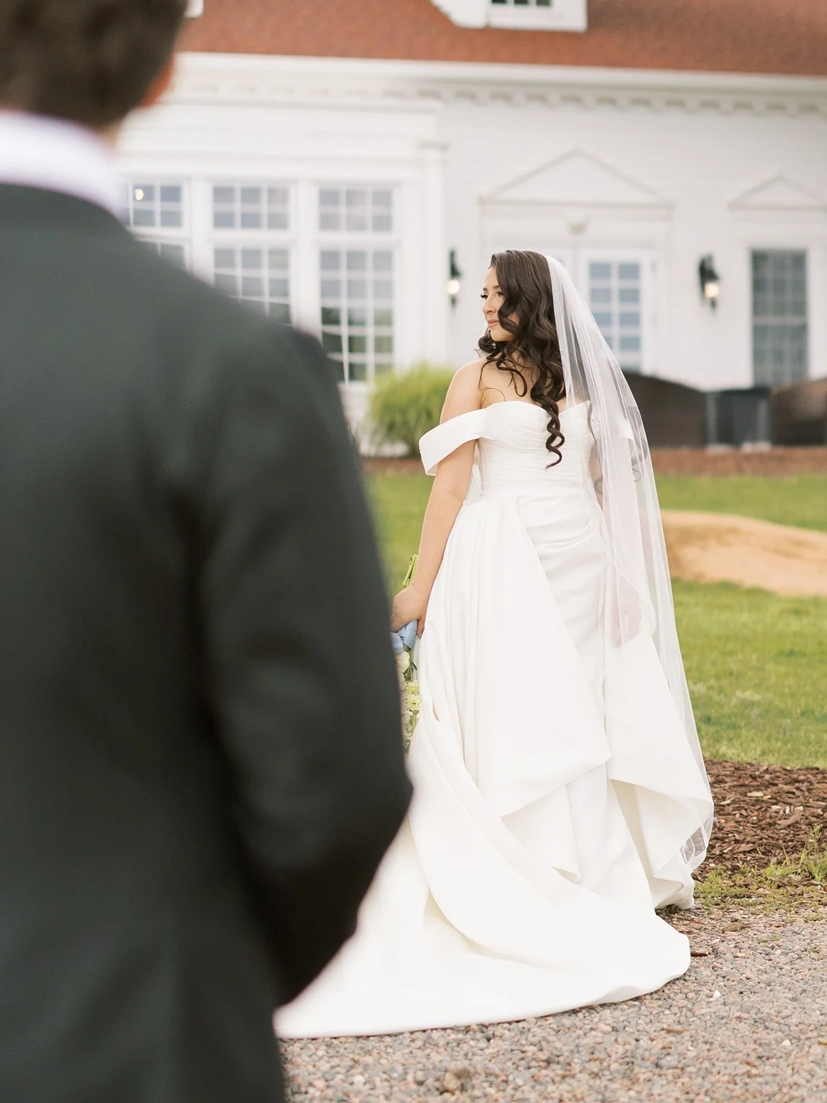 A bride in a white wedding gown with off-the-shoulder sleeves and a long veil, looking over her shoulder outside a house with white siding and large windows. A person in a dark suit is visible in the foreground, blurred.
