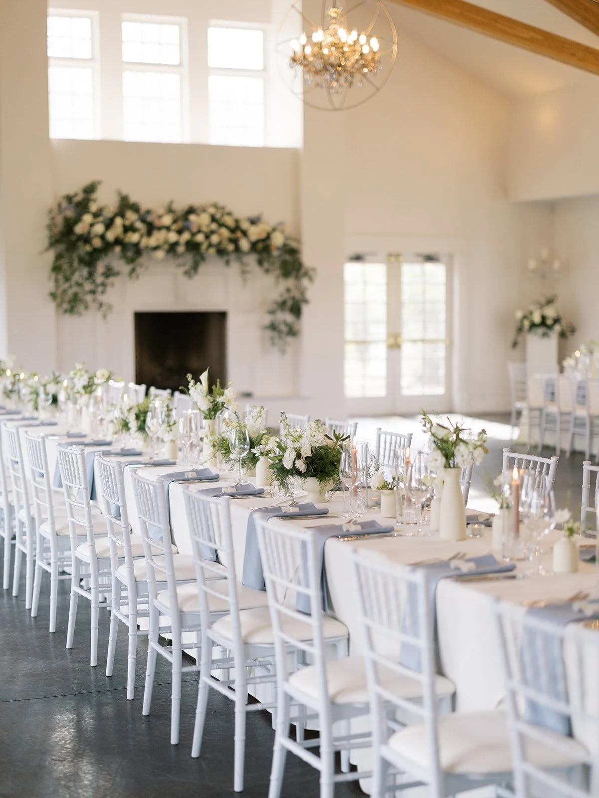 Elegant banquet table set with white tablecloths, grey napkins, floral centerpieces, and tall candles in a bright room with large windows, a fireplace with floral arrangements, and hanging chandeliers.