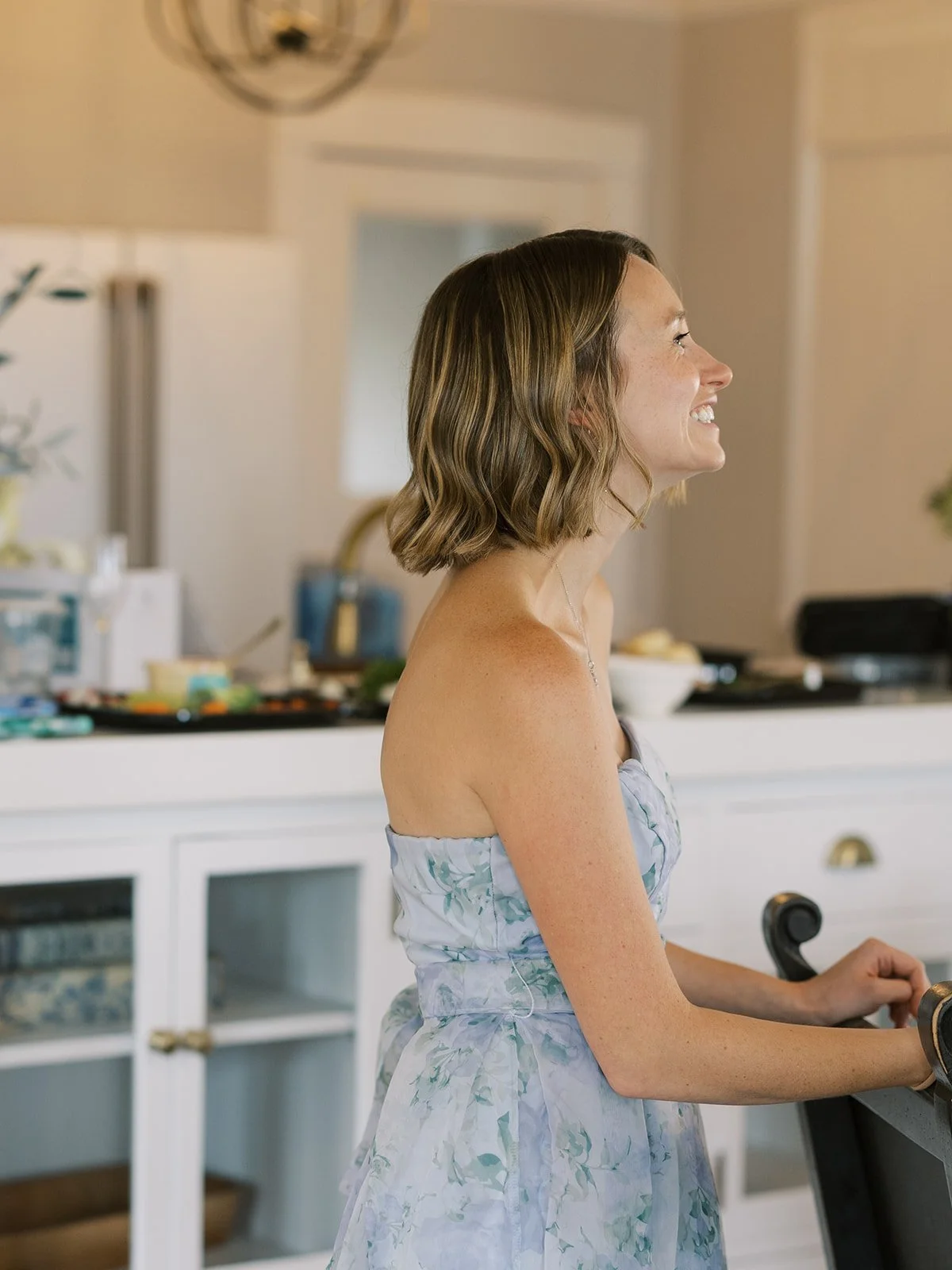 A woman with shoulder-length wavy brown hair, smiling, wearing a strapless floral dress, in a kitchen.