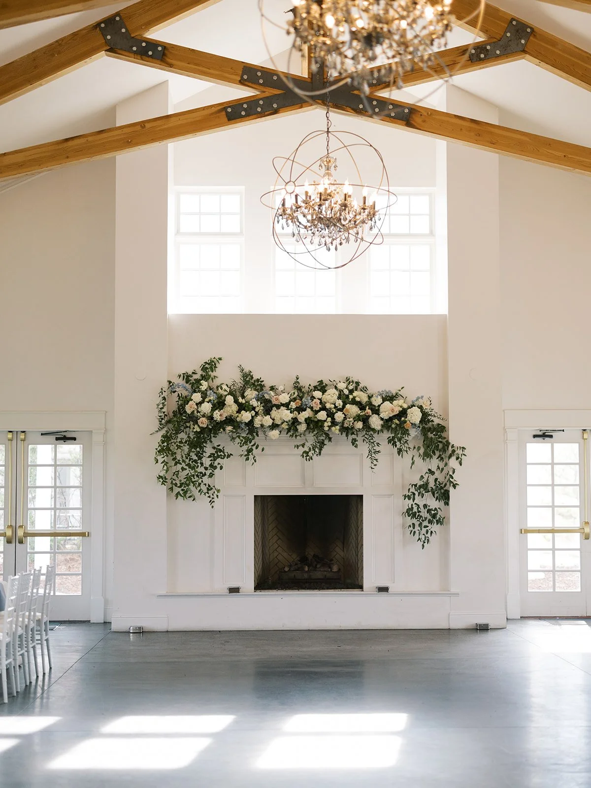 A bright, modern interior with a white fireplace decorated with a green and white floral arrangement, two windows above it, hanging chandeliers, and wooden ceiling beams.