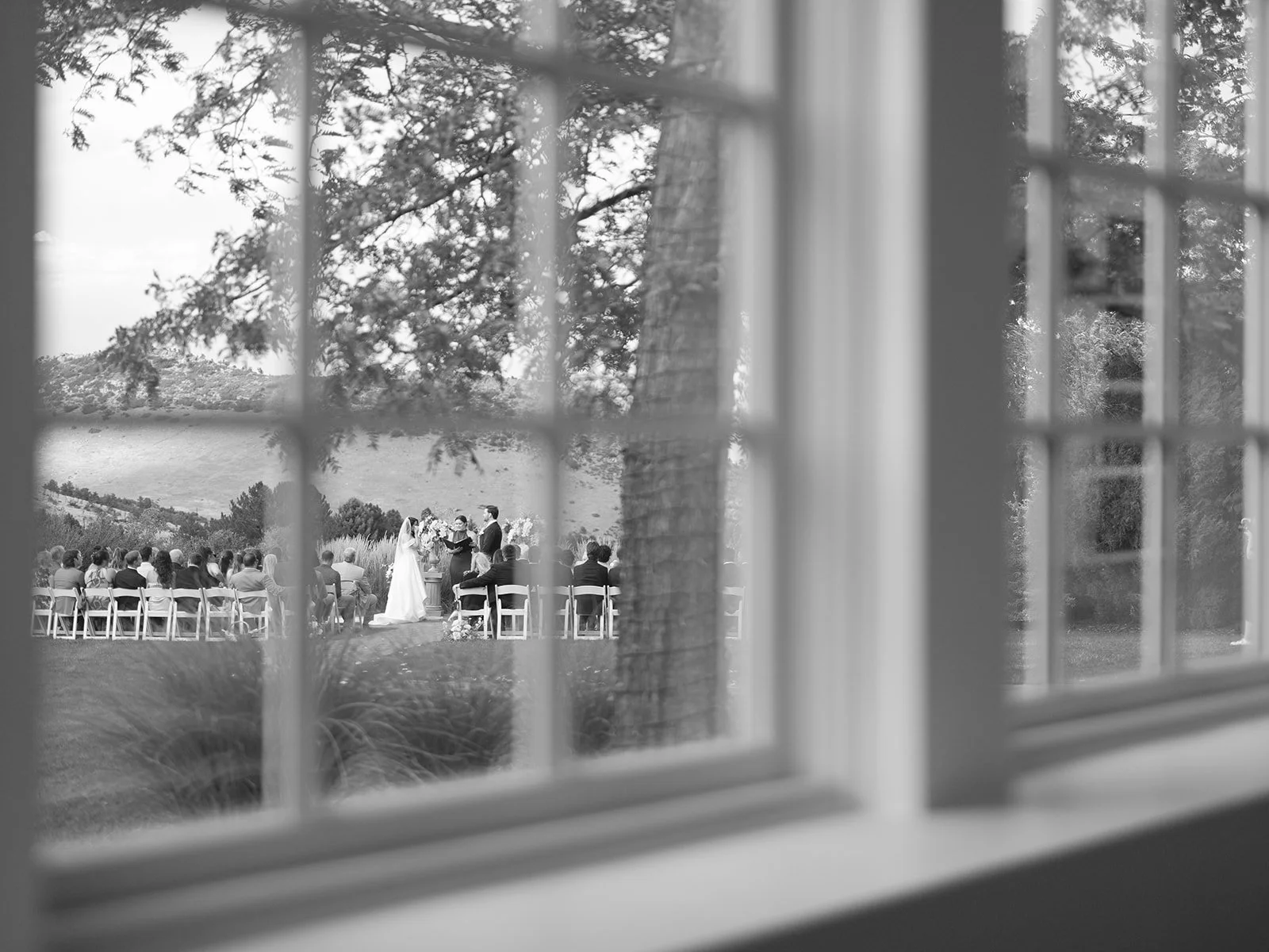 A wedding ceremony viewed through a window from inside a house, with guests seated outside on chairs and the bride and groom standing at the altar in a scenic outdoor setting.