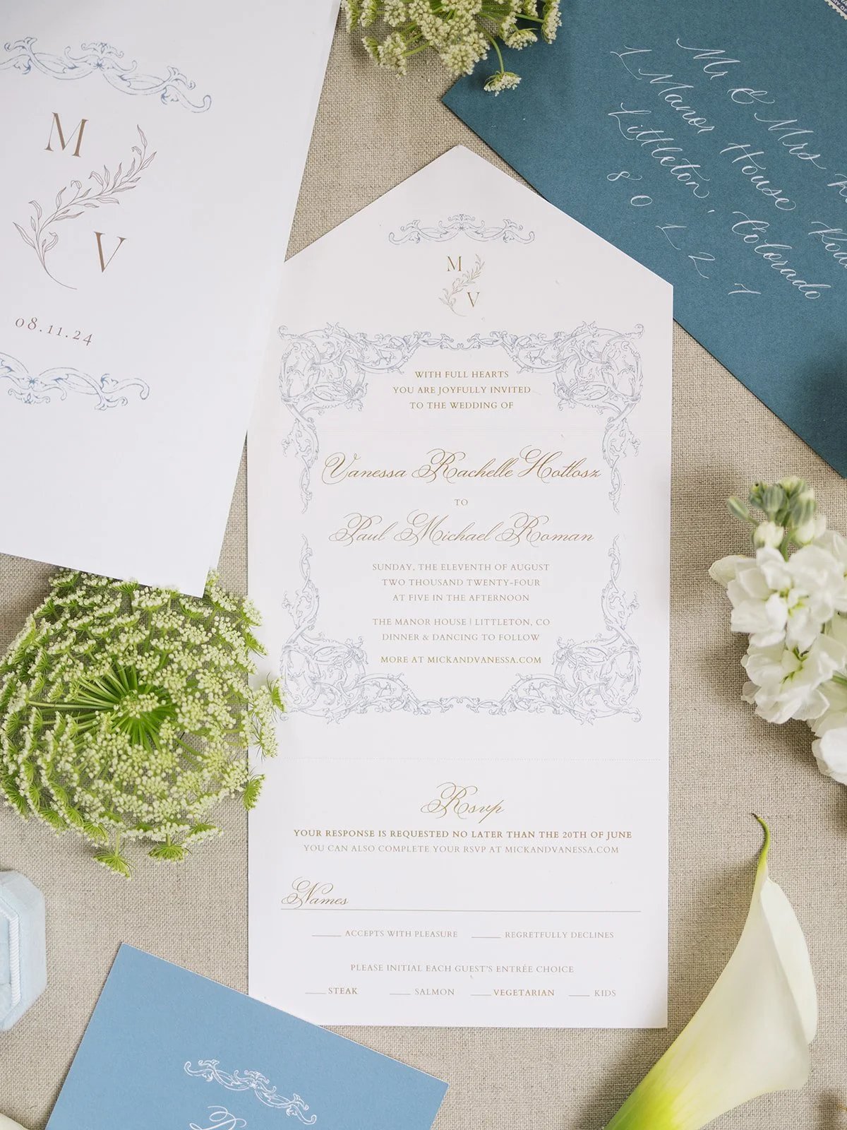 Wedding invitation with ornate border and elegant script, floral accents, surrounded by flowers, calligraphy envelopes, and decorative paper on a beige surface.