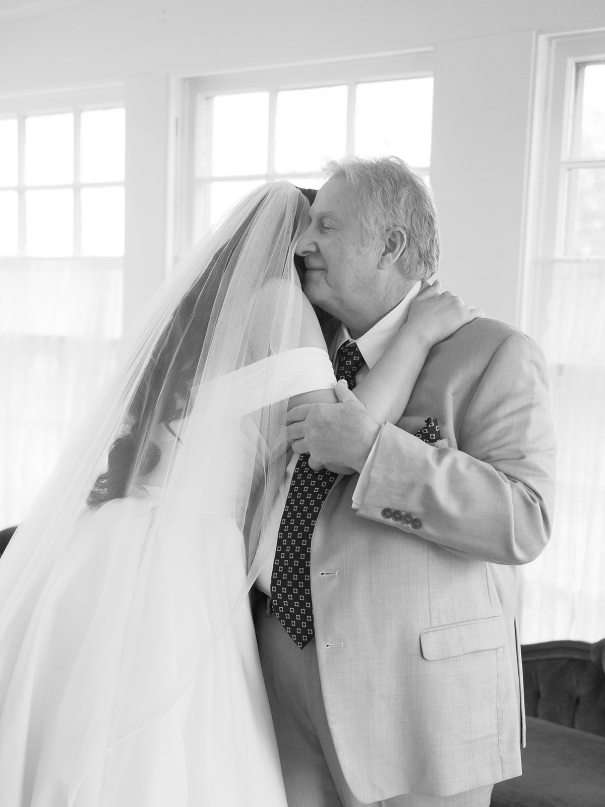 A bride and an older man, likely her father, sharing a tender moment on her wedding day inside a room with large windows, embracing each other with their eyes closed.