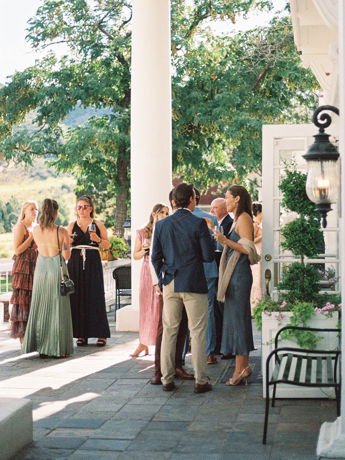 People gathering and socializing on a porch during cocktail hour, with trees and outdoor scenery visible in the background.
