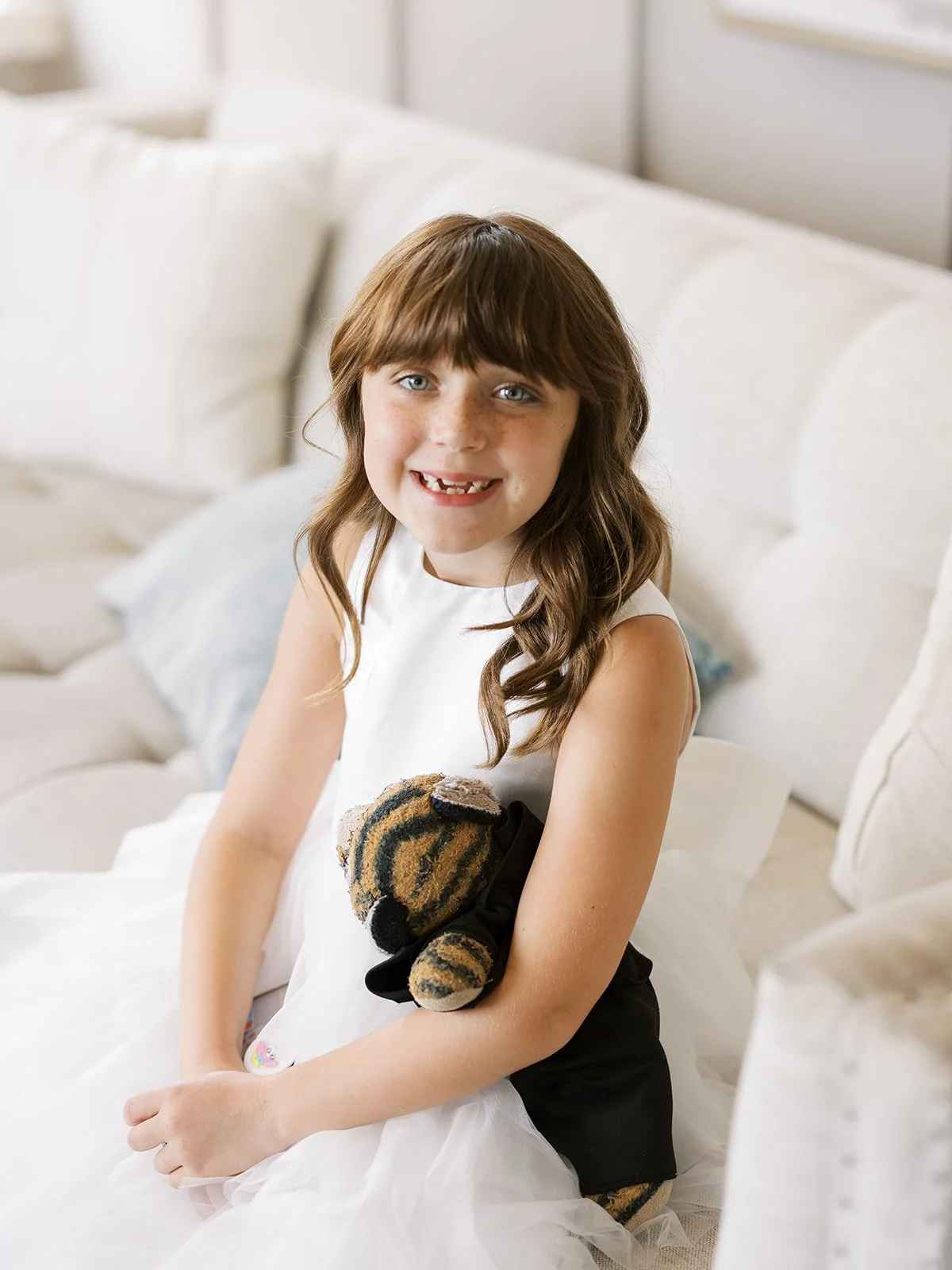 A young girl with long brown hair and freckles, wearing a white dress, sitting on a white sofa, holding a stuffed tiger toy, smiling at the camera.