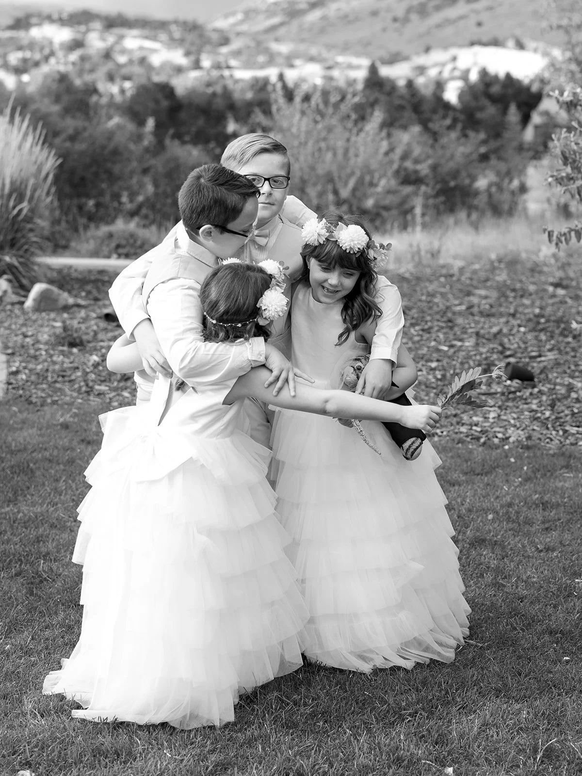 Group of children dressed in formal or costume attire, hugging and playing outdoors on grass, with a scenic background of trees and hills.