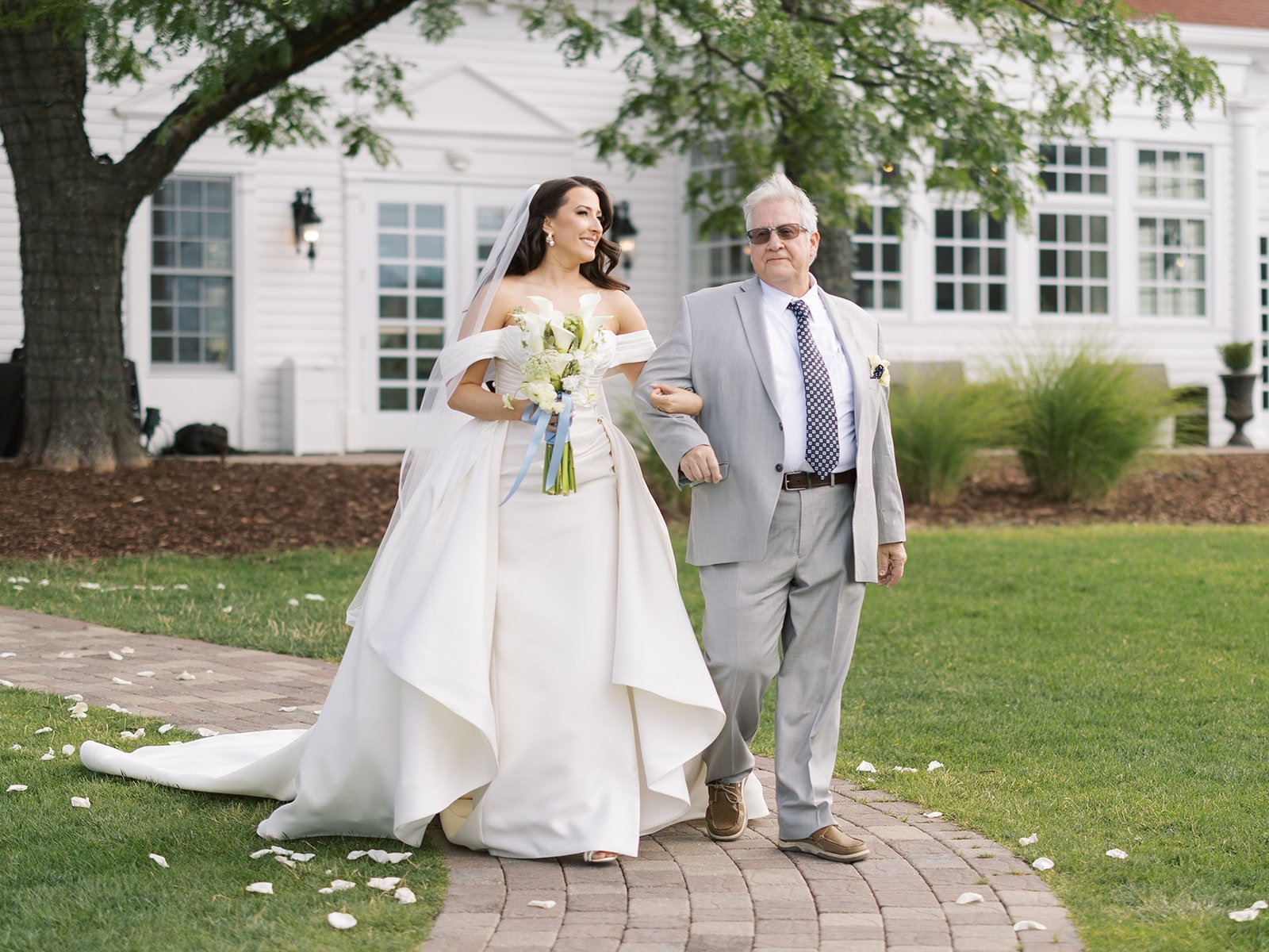 A bride in a white wedding dress holding a bouquet walking with an older man in a light gray suit outside a white house. The bride is smiling and the man is walking with a serious expression. They are on a brick pathway surrounded by grass, with scat