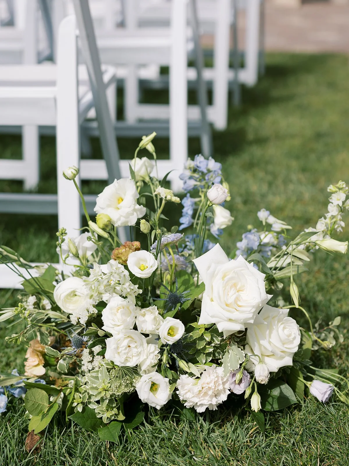 A white floral arrangement on grass with white chairs in the background.