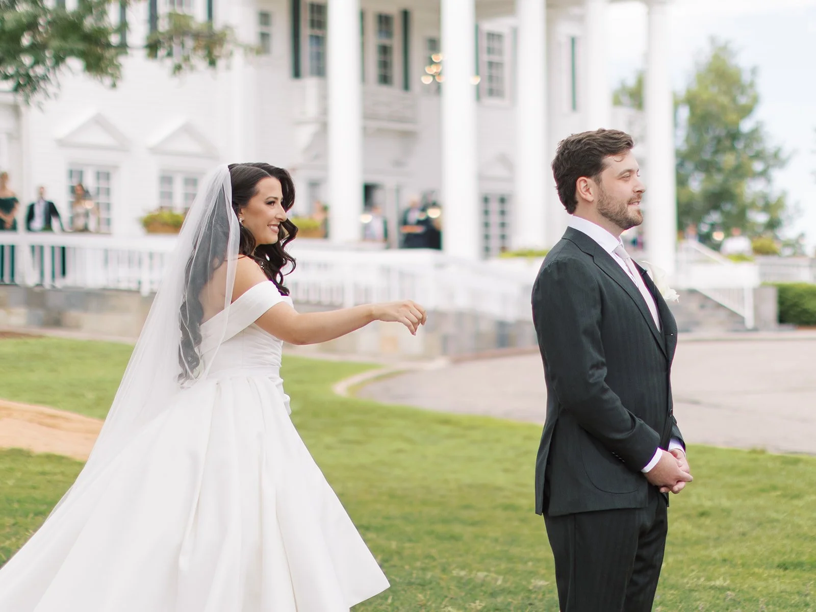 Bride in a white wedding dress and veil reaching out towards groom in a black suit on a lawn outside a white building with columns.