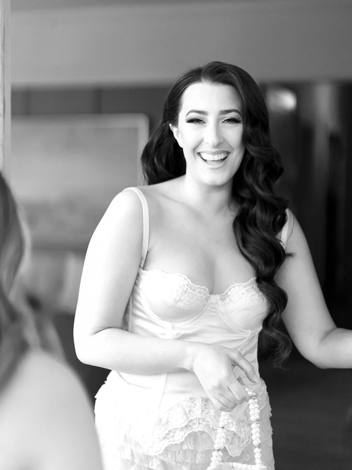 A woman with long dark wavy hair smiling and holding a string of pearls, wearing a white lace-trimmed top.