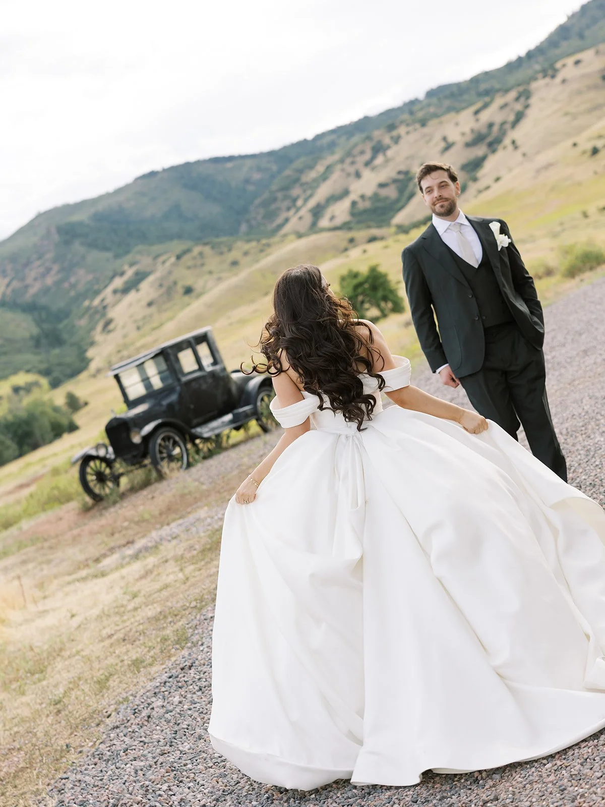 A bride in a white wedding gown with long dark hair and a groom in a black suit and tie standing outdoors on a gravel path with hills and an old black car in the background.