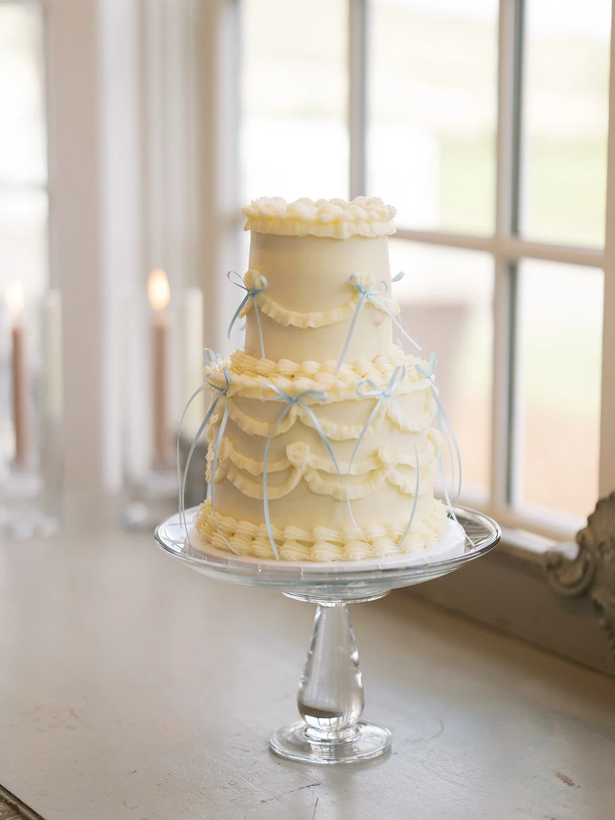 A three-tier wedding cake with white icing, decorated with blue ribbons and bow ties, and piped with white frosting, placed on a glass cake stand near a window.