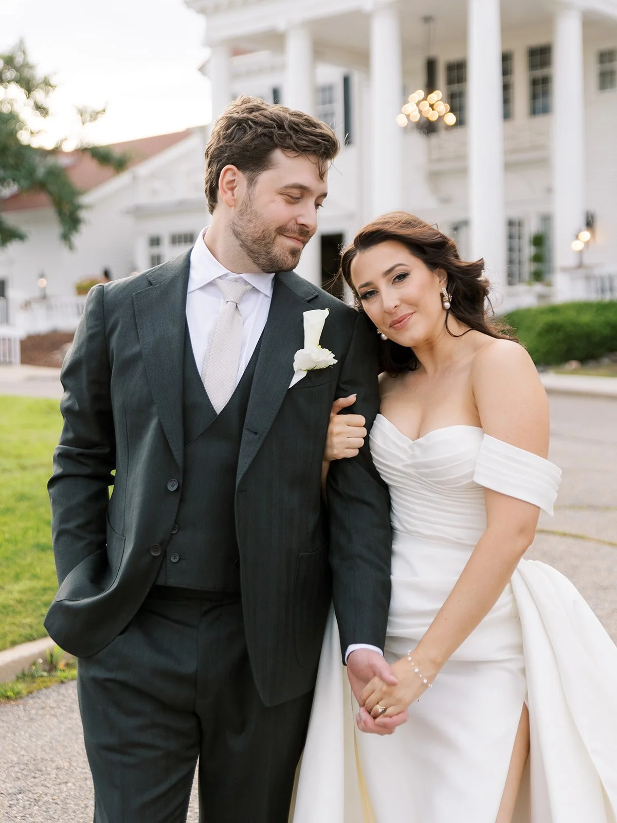 A bride and groom standing outside a large white wedding venue, holding hands and smiling. The groom is wearing a dark suit with a white shirt and tie, and has a white boutonniere. The bride is in a white off-shoulder wedding gown with her hair style