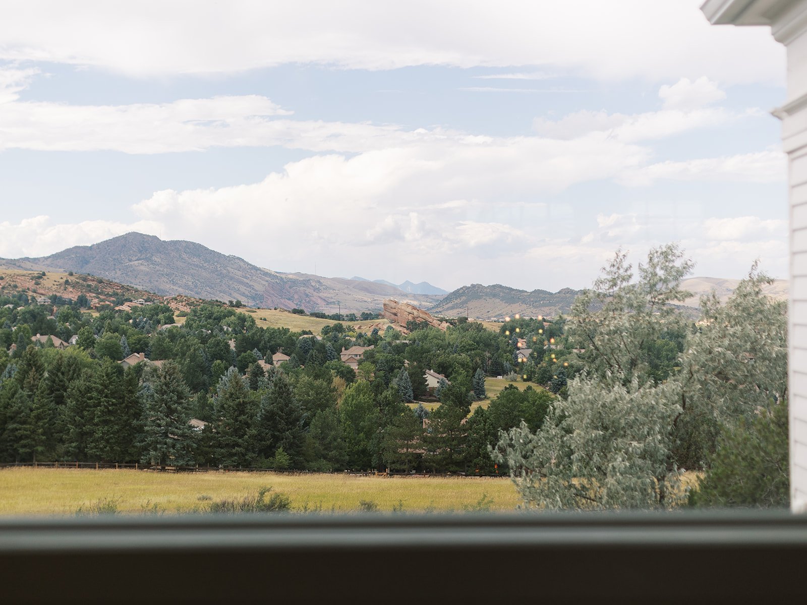 View from a window overlooking a rural landscape with trees, houses, and mountains under a partly cloudy sky.