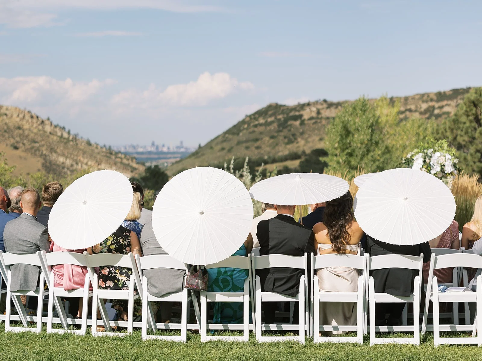 Wedding ceremony outdoors with guests seated in white chairs, some holding white parasols. The setting is in a scenic area with hills and a city skyline in the background.