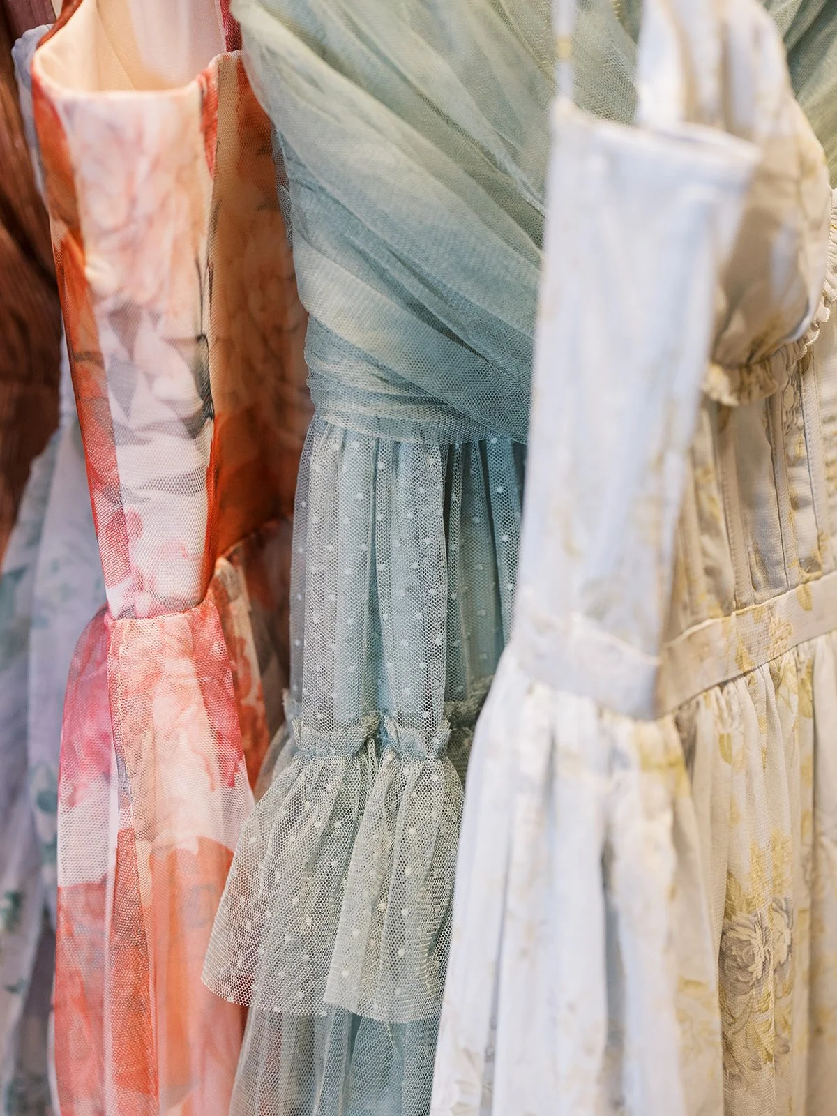 Close-up of three dresses with floral and pastel patterns, ruched and pleated fabric, hanging on a rack.