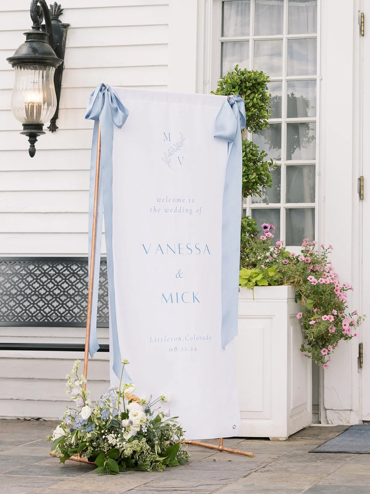 Wedding welcome sign for Vanessa and Mick with floral arrangement and greenery outside a house.