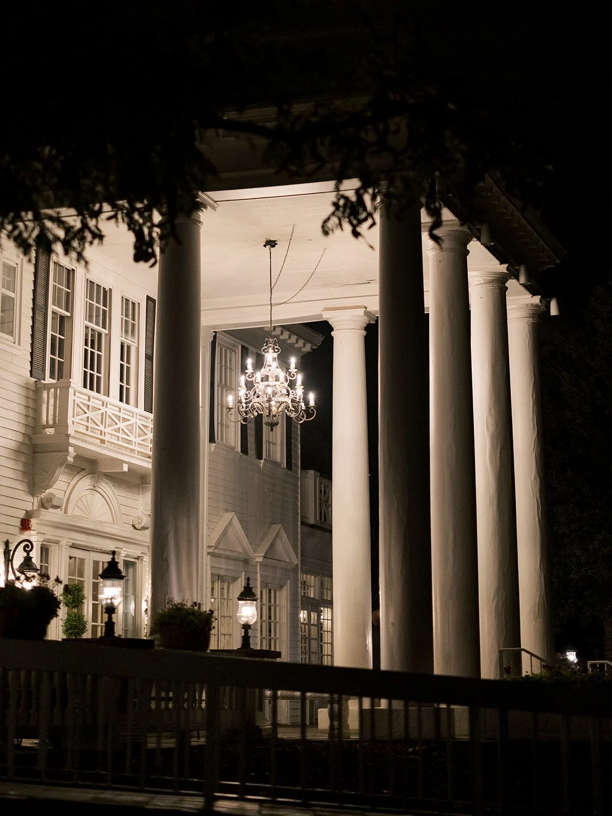 Night view of a large, white, classical-style house with tall columns, a chandelier hanging from the ceiling, lamps on the porch, and potted plants.