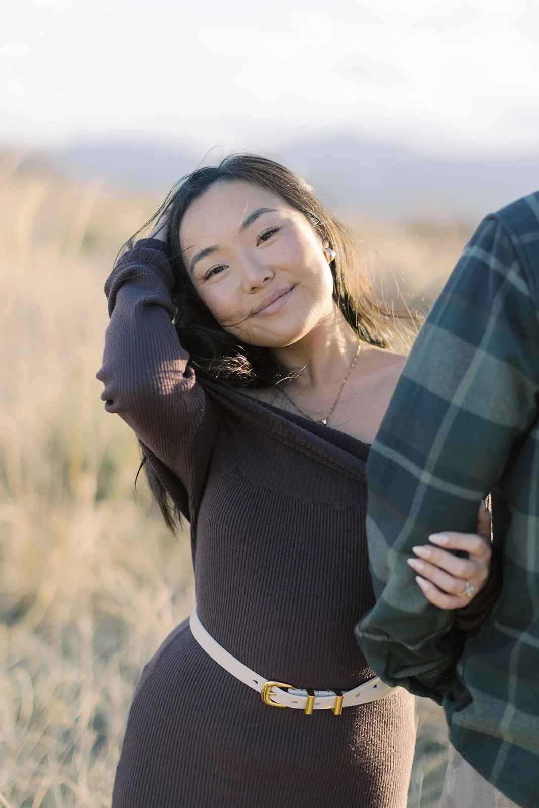 A woman with long dark hair smiling at the camera while standing outdoors in a field of tall grass, wearing a dark brown dress and white belt.