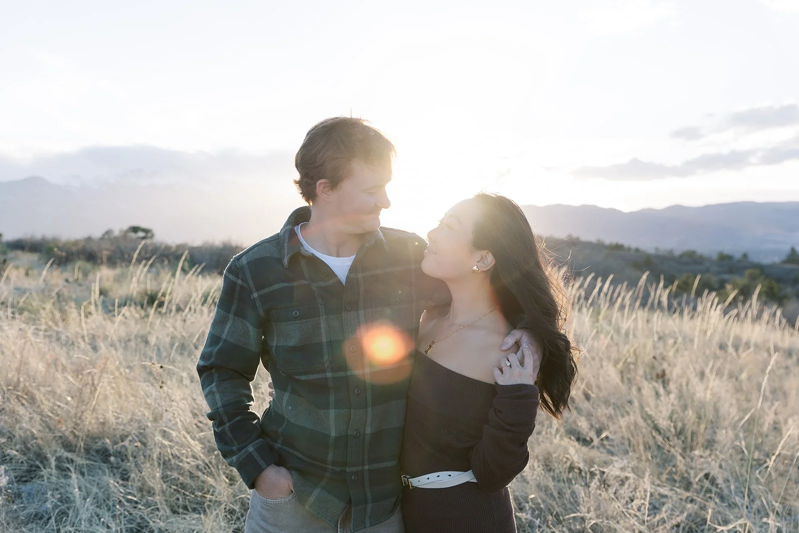 A couple standing close together in a field at sunset, looking into each other's eyes.