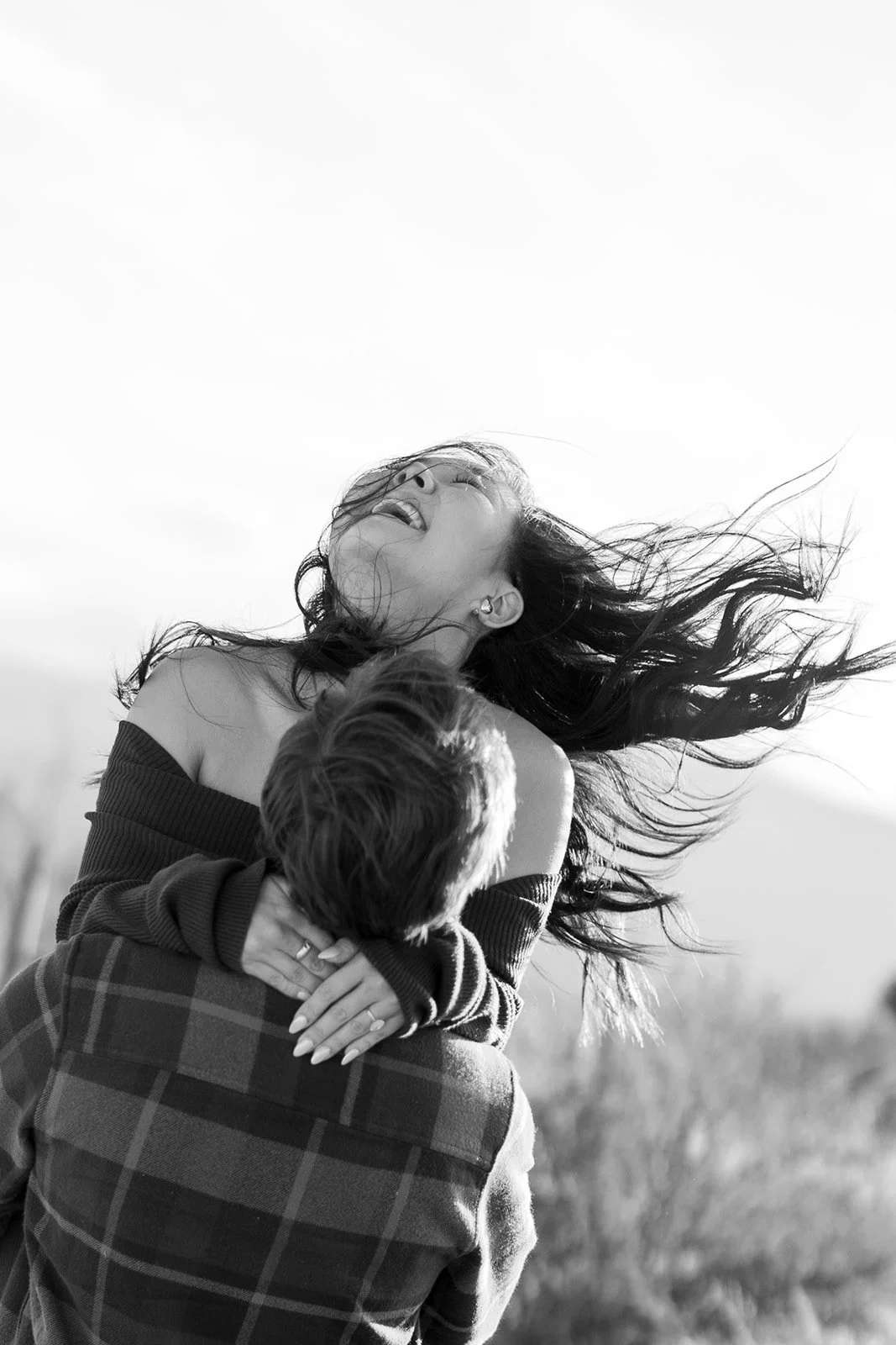 A woman with long flowing hair embraces a young boy outdoors, both appear joyful, in a black and white photograph.