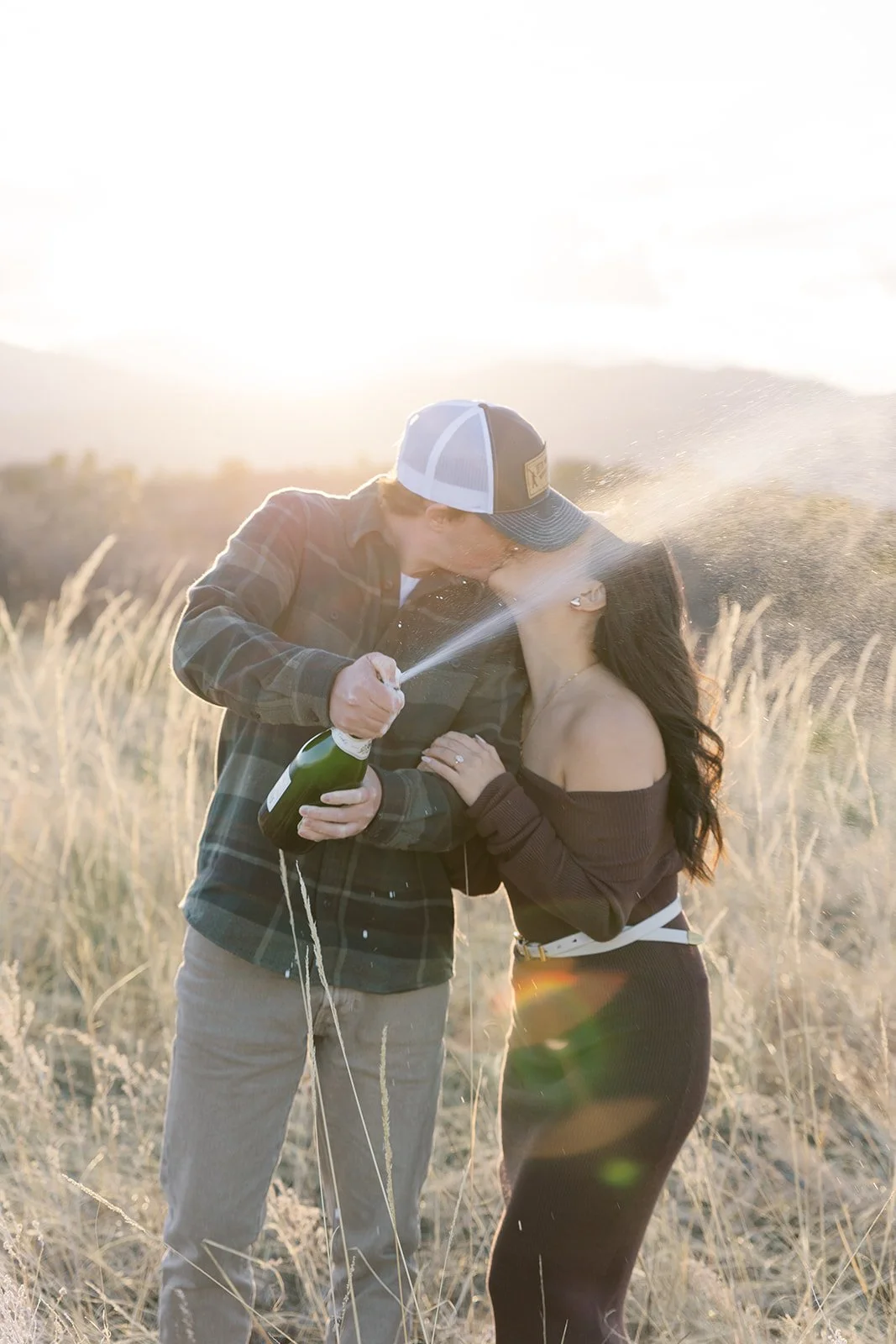 A couple kissing outdoors in a field at sunset, with the man spraying champagne, creating a celebratory scene.