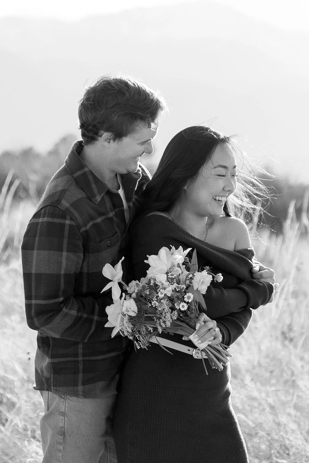 A young man and woman share a happy moment outdoors, with the man holding a bouquet of flowers, both smiling and laughing in a field.